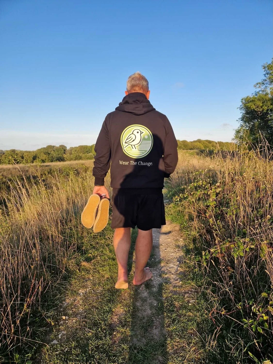 Man walking barefoot on a dirt trail through a grassy field, holding shoes in his left hand, wearing a black hoodie with a bird logo and the text 'Wear The Change', during daytime with clear blue sky.