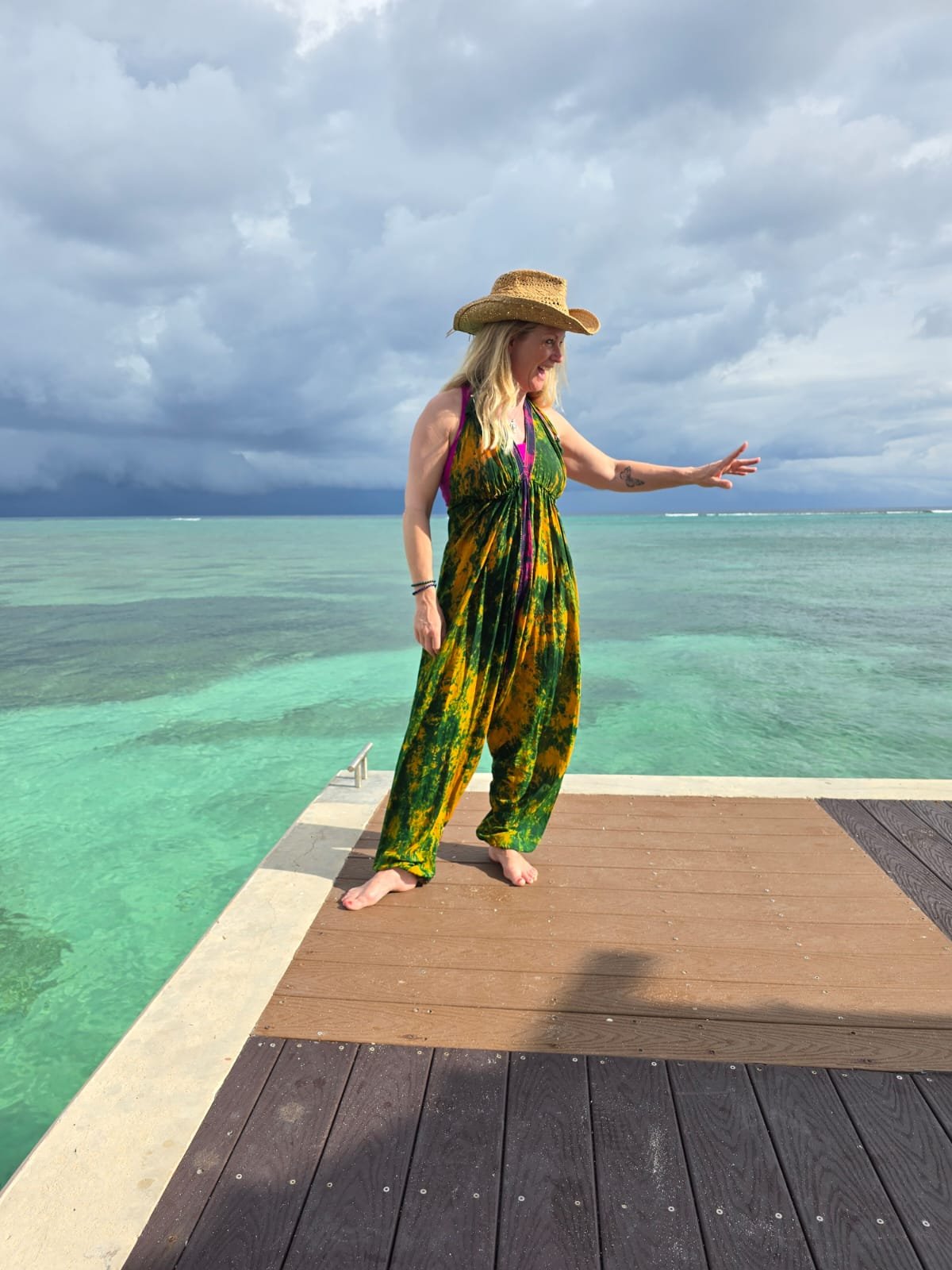 Woman in colorful dress and straw hat standing on dock by ocean during cloudy weather, with storm clouds in the background.