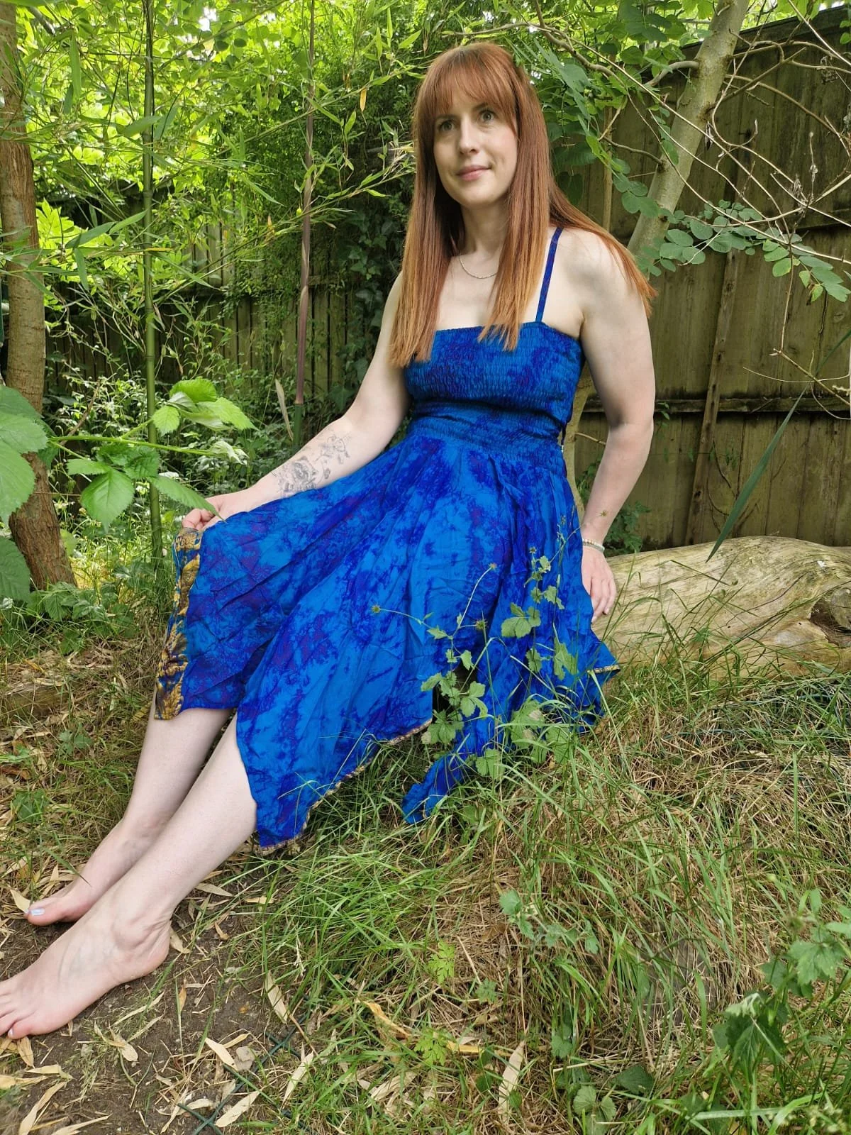 A woman with red hair in a blue dress sitting outdoors on a grassy area surrounded by green trees and plants.