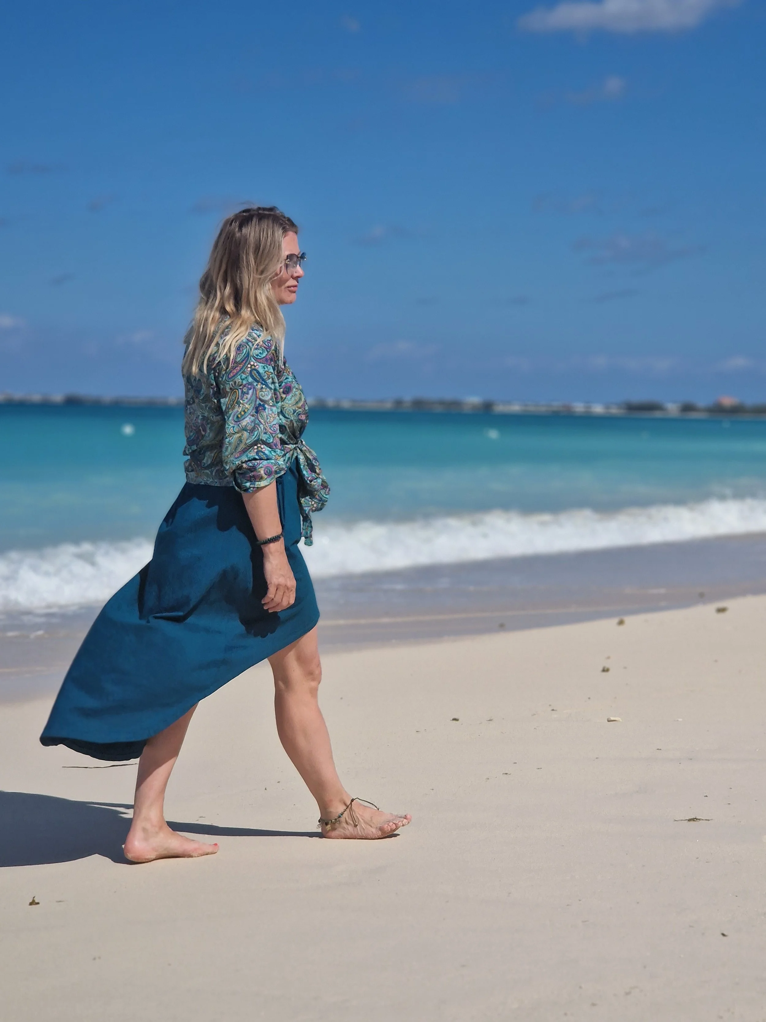 A woman walking barefoot on a sandy beach near the ocean, with blue water and a partly cloudy sky in the background.