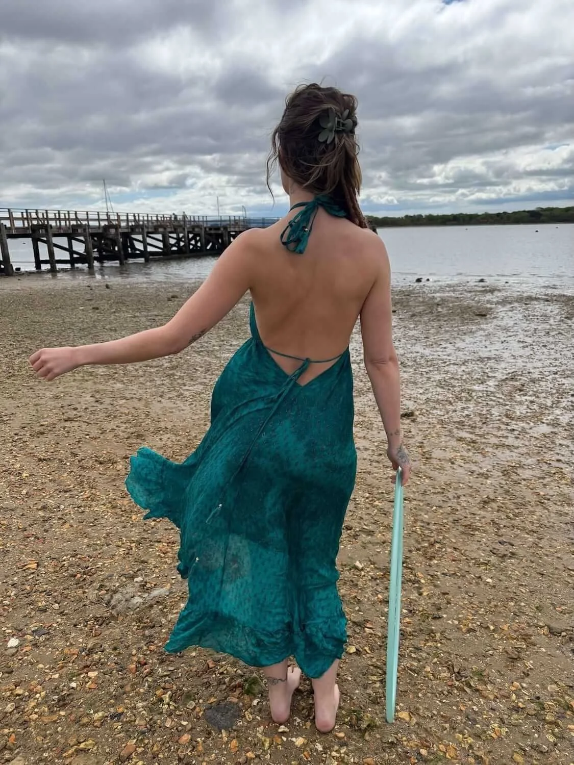 Woman in a teal dress walking barefoot on a pebbled beach towards water with a wooden dock in the background and cloudy sky.