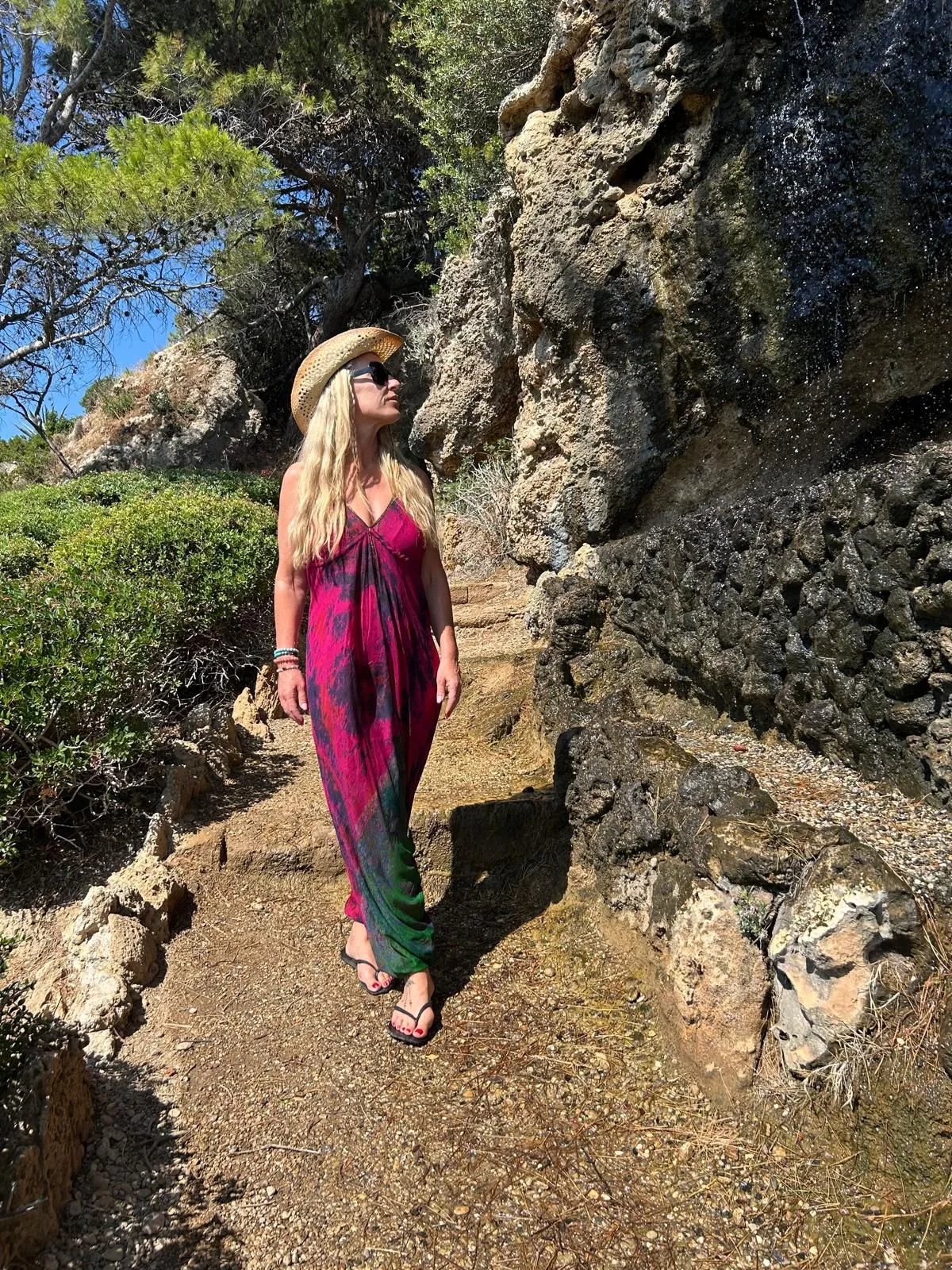 Woman in a pink and green dress, wearing a straw hat and sunglasses, walking on a rocky trail near a large rock formation, with green bushes and trees around.