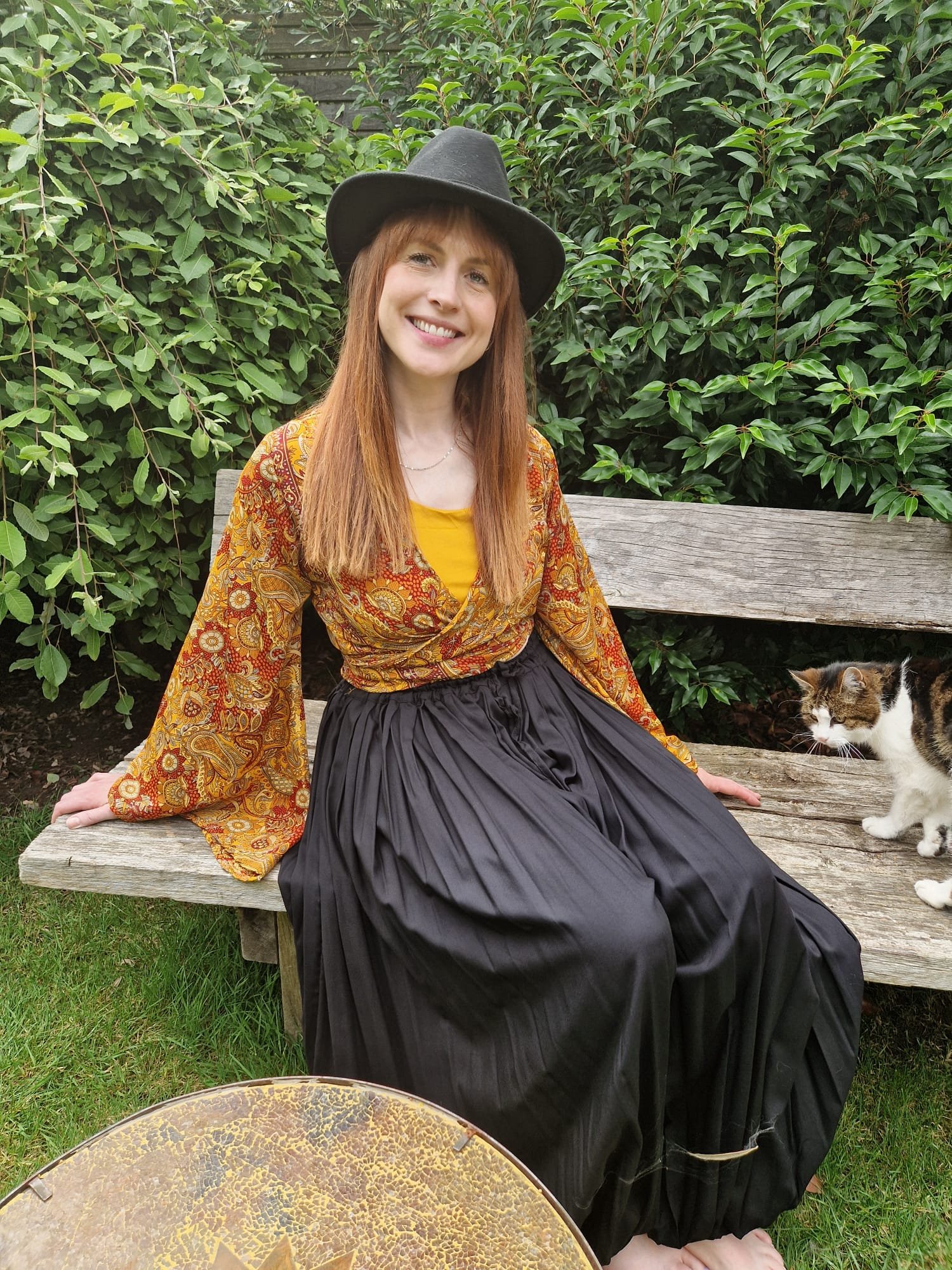 A woman with long red hair wearing a black wide-brimmed hat, a yellow top, a patterned orange and yellow coat, and a black pleated skirt, is sitting on a wooden bench outdoors. She is smiling and looking at the camera, with bushes in the background a