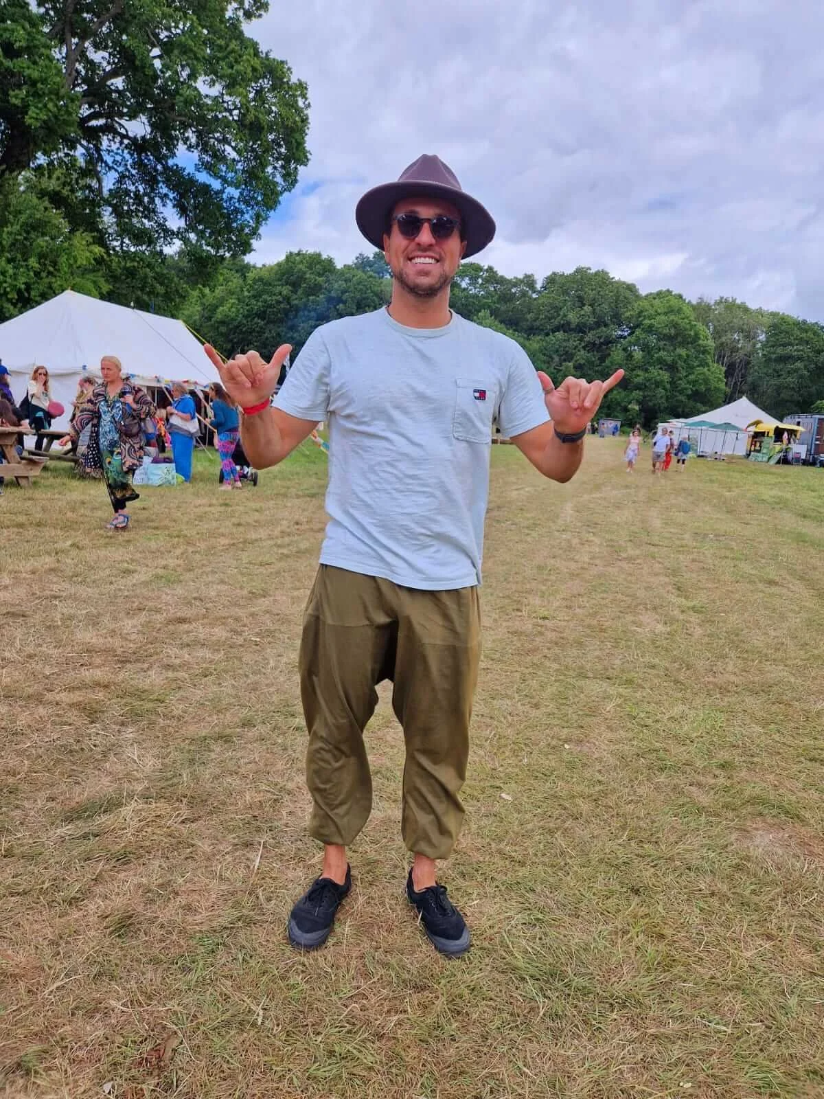 A man smiling at an outdoor event wearing a gray T-shirt, khaki pants, sunglasses, a wide-brimmed hat, and black shoes. He is making a shaka sign with both hands. In the background, there are tents, trees, and other people.