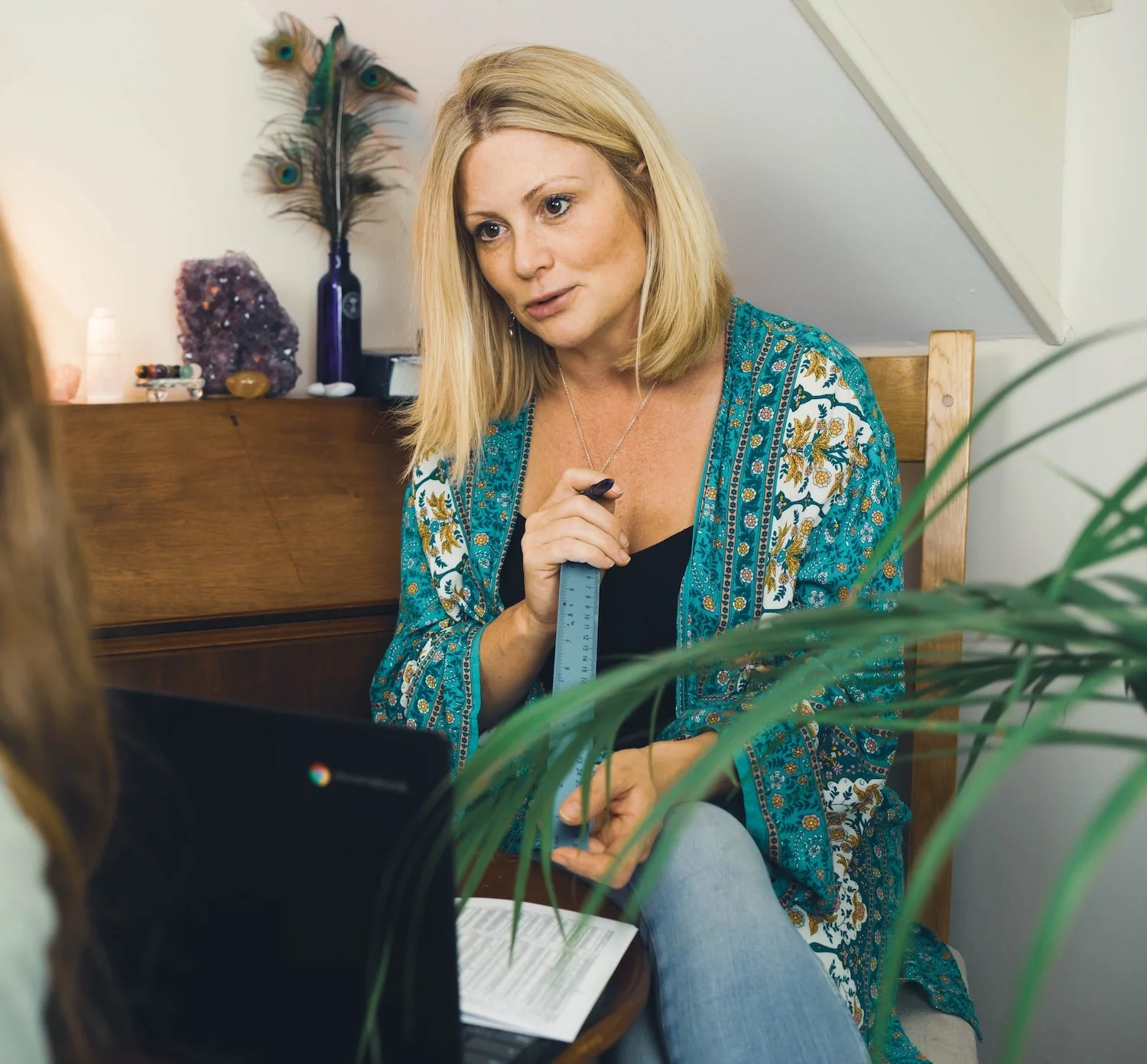 A woman with blonde hair and a blue patterned cardigan sitting in front of a webcam, holding a ruler, engaged in a conversation during a video call. There are decorative items, including feathers and crystals, on a table behind her.