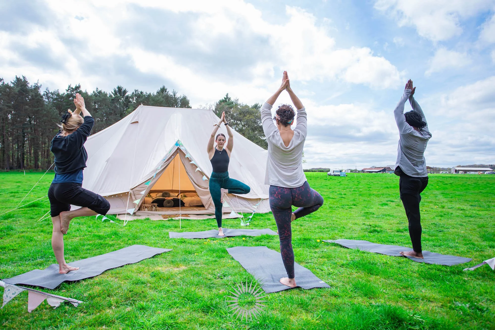 Four women practicing yoga outdoors on green grass, with a large white tent and trees in the background, under a partly cloudy sky.