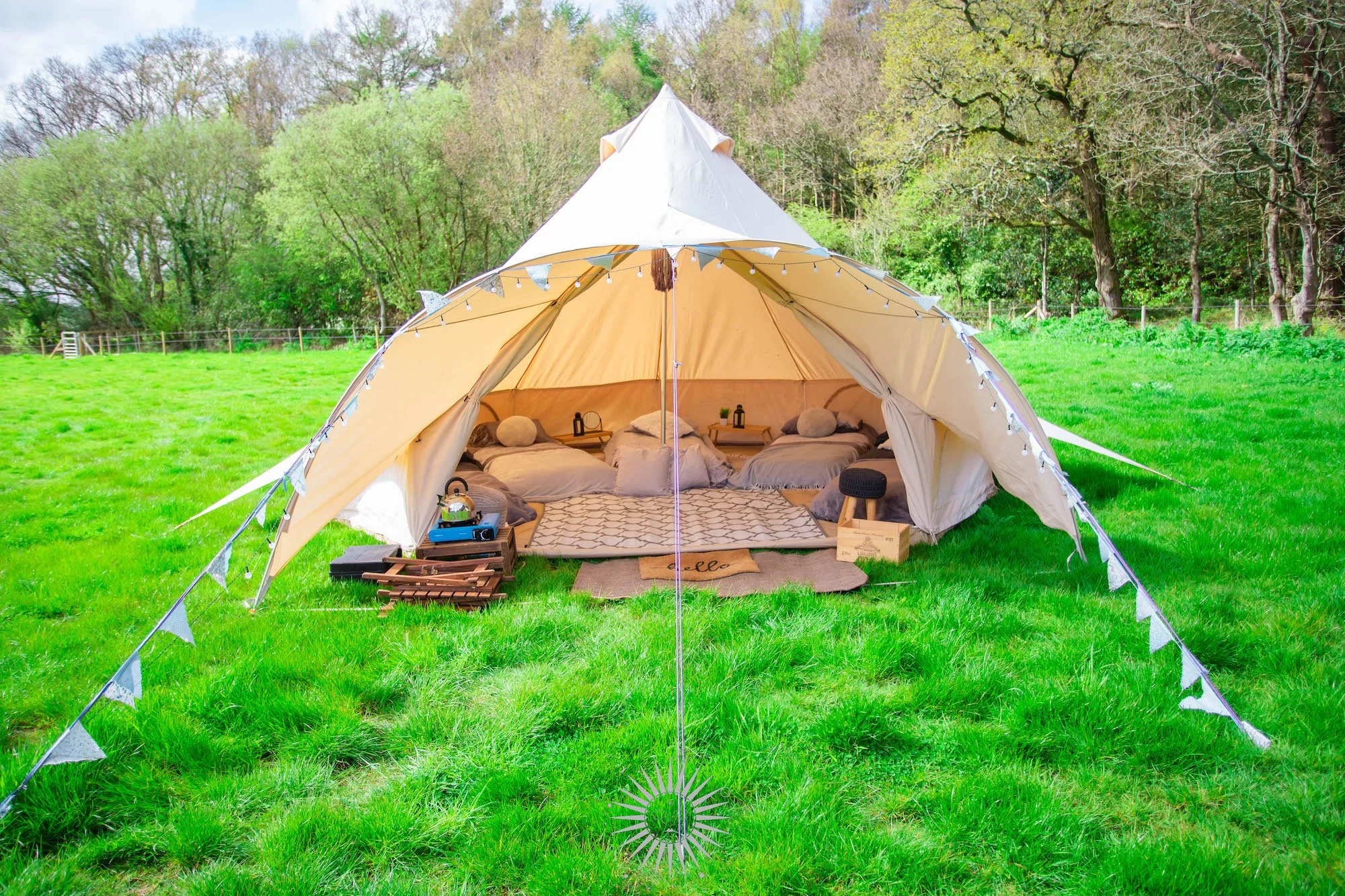 A cozy camping tent set up in a lush green field with trees in the background, decorated with string lights, and featuring beds and camping gear inside.