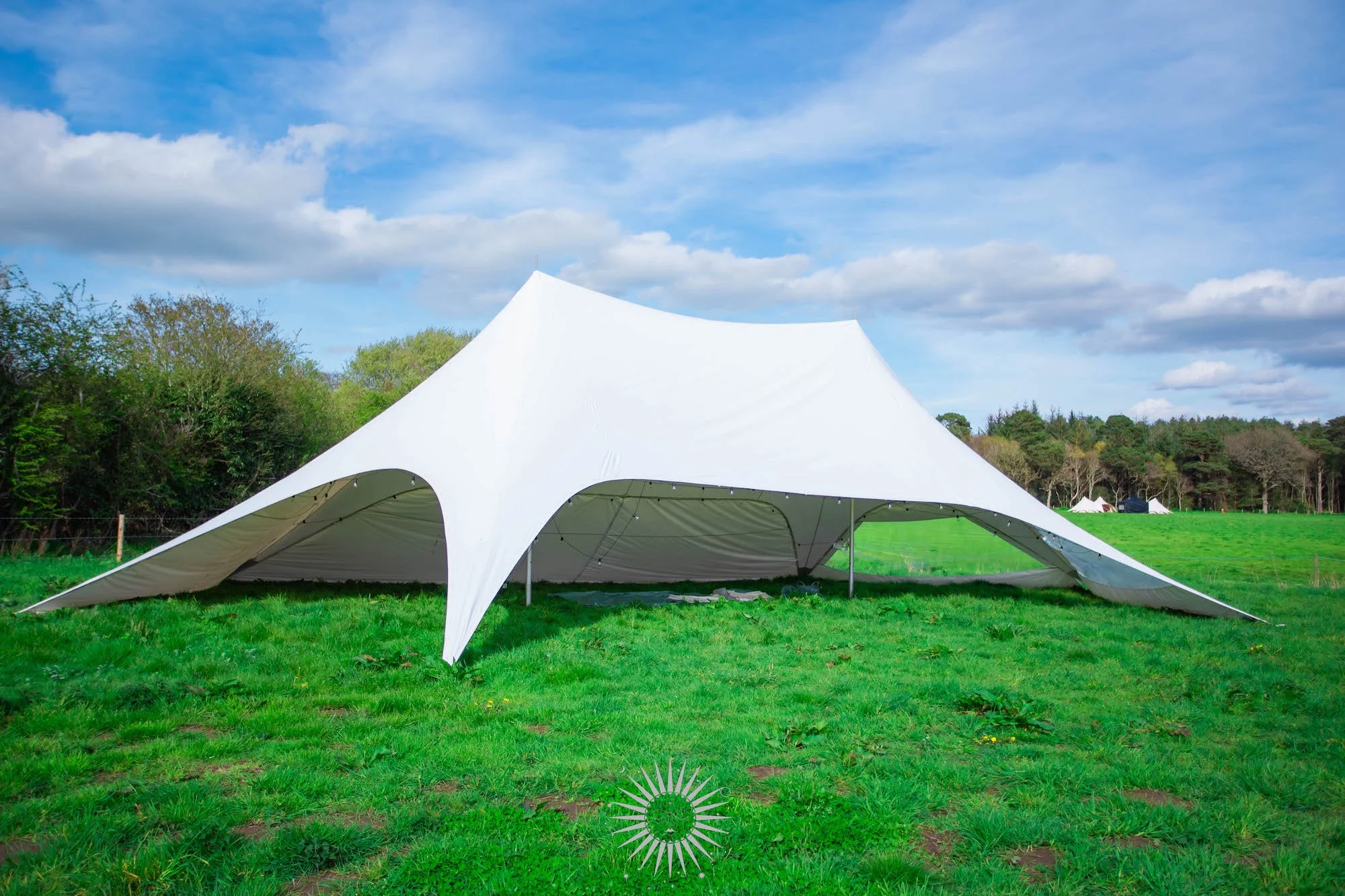 A large white tensile tent set up on a grassy field with trees in the background and a cloudy blue sky overhead.