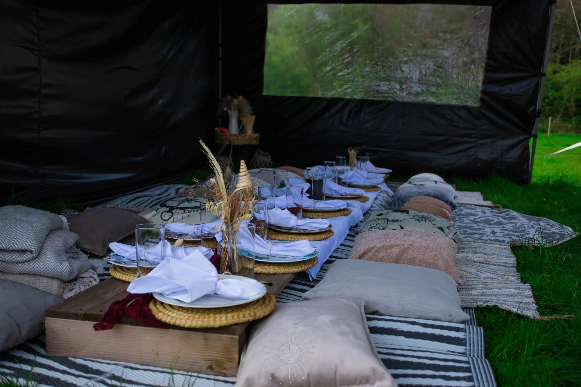 A cozy outdoor picnic setup on a striped blanket with cushions, featuring a low table with white napkins, glasses, and decorative dried flowers, inside a black tent on a grassy area.