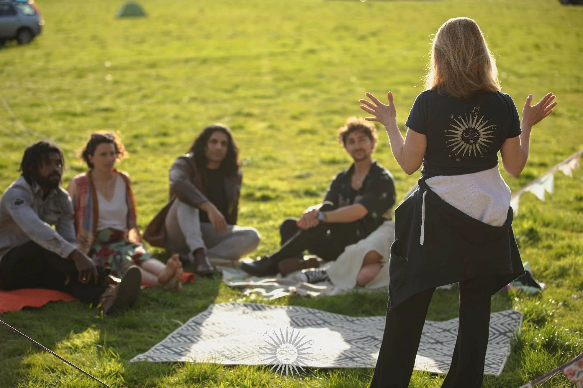 A woman standing outdoors appears to be talking to a group of four people sitting on blankets on a grassy field during a sunny day.