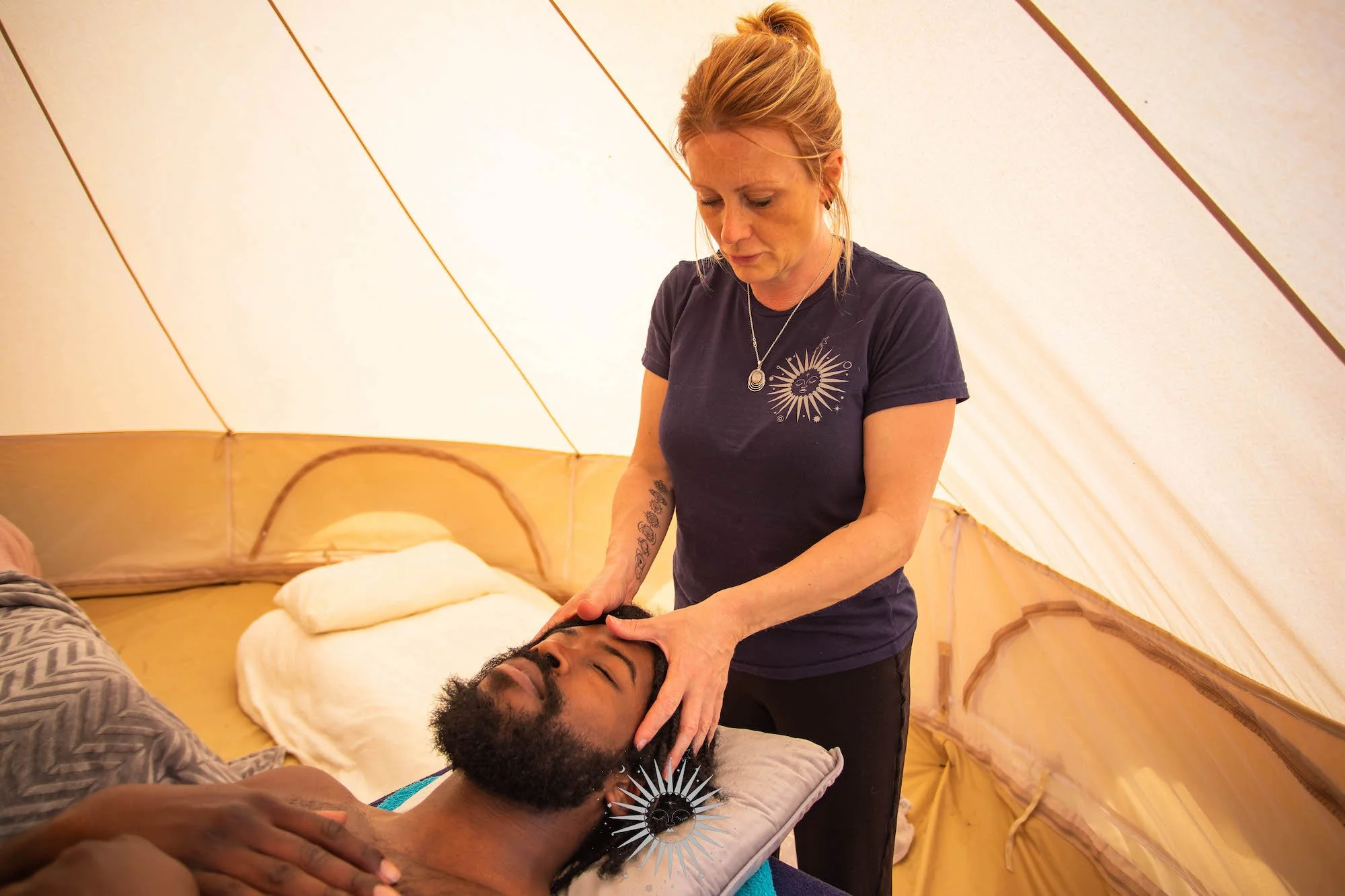 A woman providing a massage or healing treatment to a man lying down inside a tent.