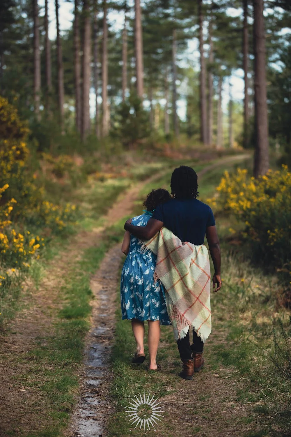 A couple walking arm in arm down a wooded trail, surrounded by yellow flowers and tall trees, with one person holding a blanket.