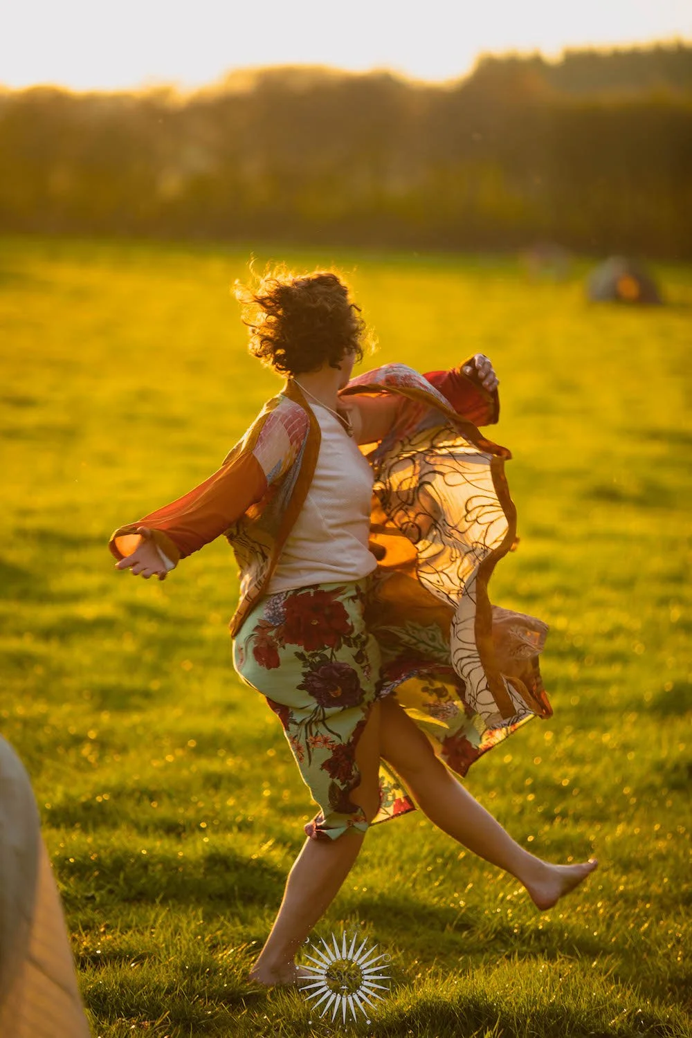 A person dancing or spinning in a grassy field during sunset, wearing colorful, floral-patterned clothing and with curly hair.
