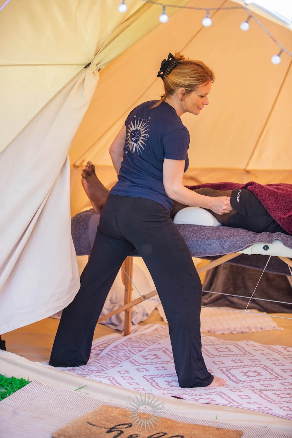 A woman receiving a massage in a cozy tent with string lights at sunset.