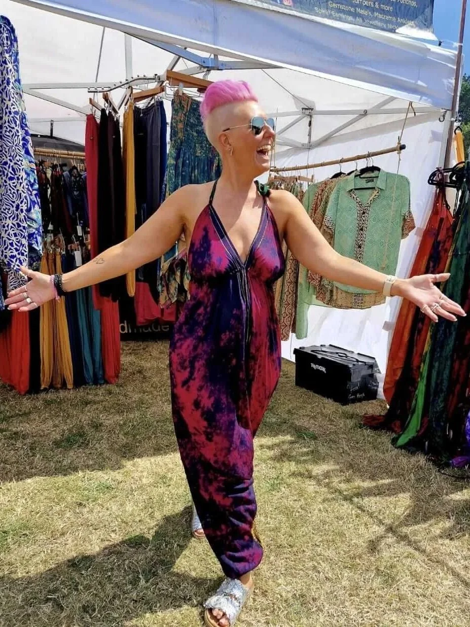 A woman with pink hair and sunglasses smiling and spreading her arms at an outdoor market stall selling colourful clothing.