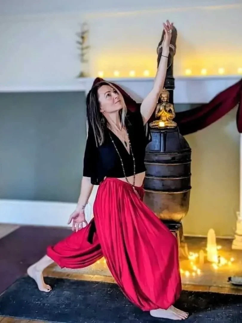 A woman in a yoga pose with one arm raised and the other hand on her head, standing near a large decorative object in a room decorated with candles and drapery.
