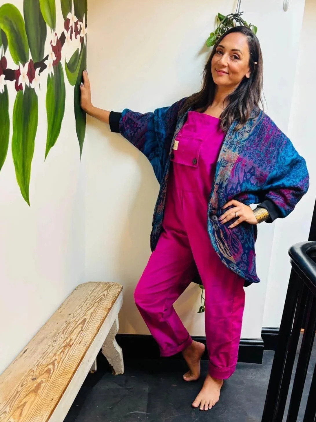 A woman in pink dungarees and blue patterned cardigan stands barefoot next to a wall with a floral mural, posing with her hand on the wall and smiling.