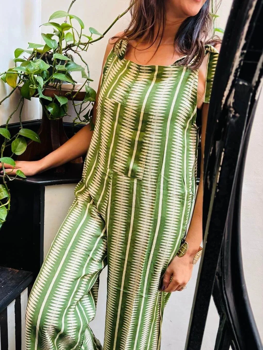 Woman taking a mirror selfie wearing a green and cream patterned jumpsuit, standing next to a potted plant.