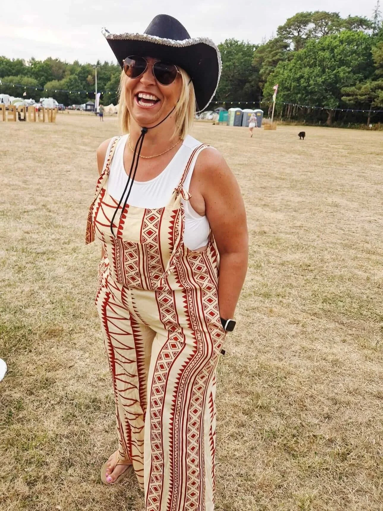 Smiling woman wearing a black cowboy hat, sunglasses, a white sleeveless top, and patterned overalls, standing on a grassy field at outdoor event with tents, portable toilets, and trees in the background.
