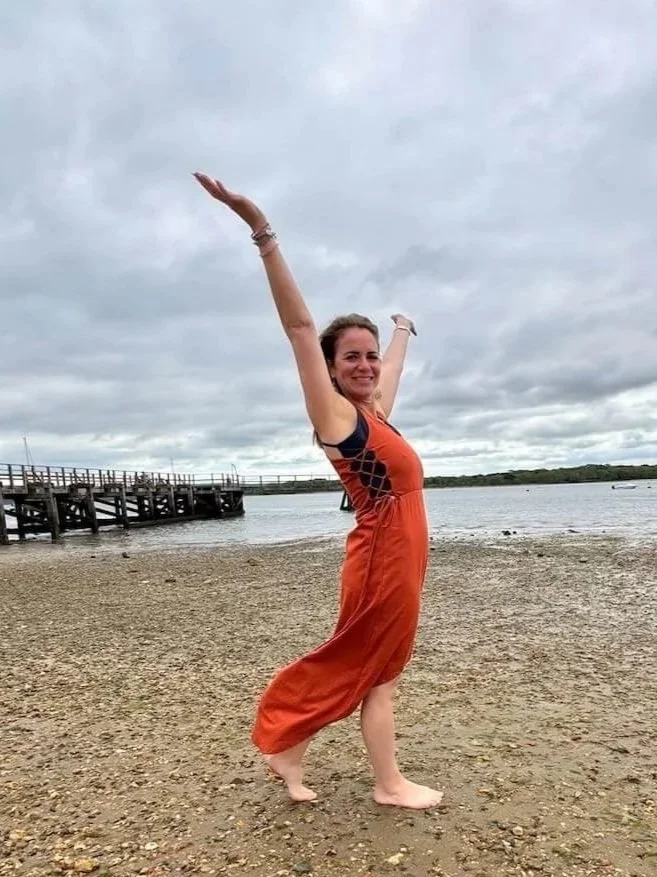 A woman in an orange and black dress standing barefoot on a pebbly beach, smiling with arms raised, near water and a cloudy sky, with a wooden pier in the background.