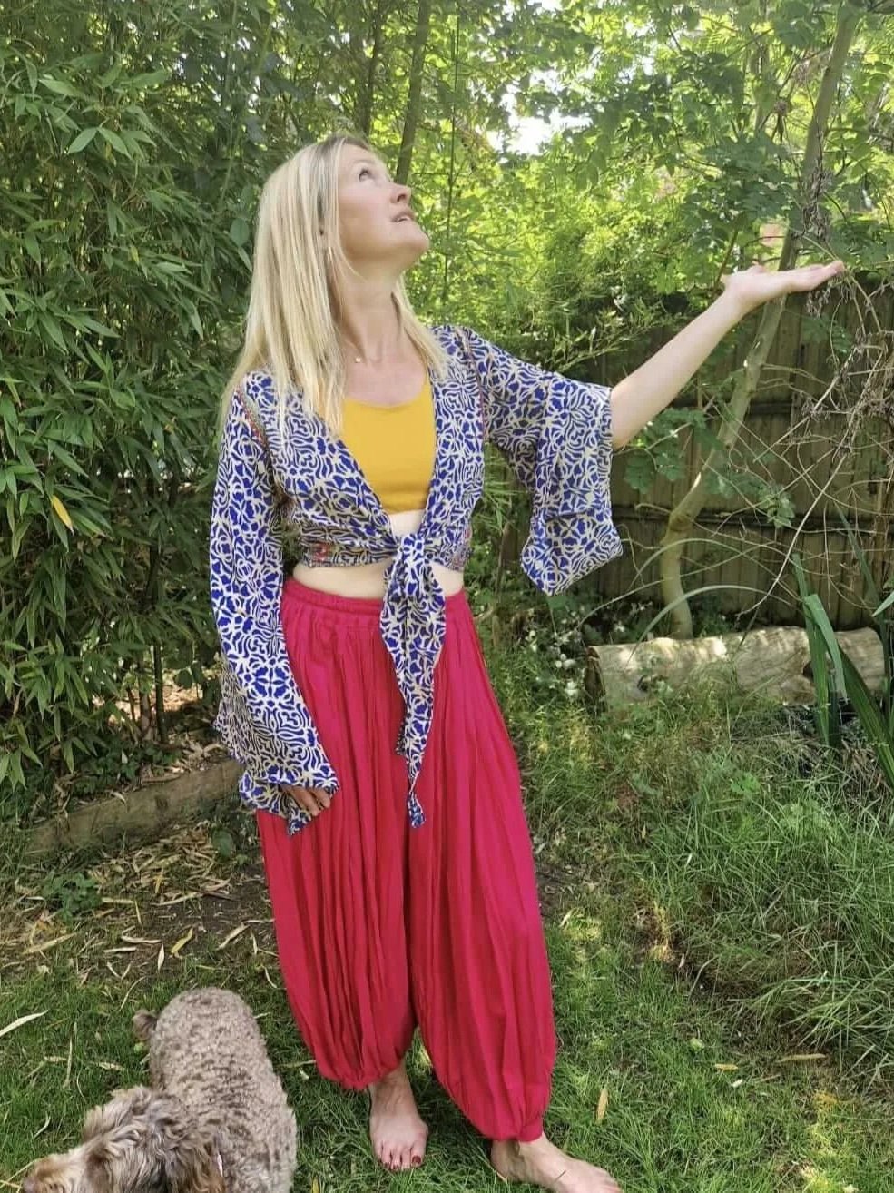 A woman standing in a lush garden, looking up and to her left, with one arm extended and hand open. She has long blonde hair and is wearing a yellow top, a blue patterned jacket tied at the waist, and bright red flowy pants. There is a small dog at h