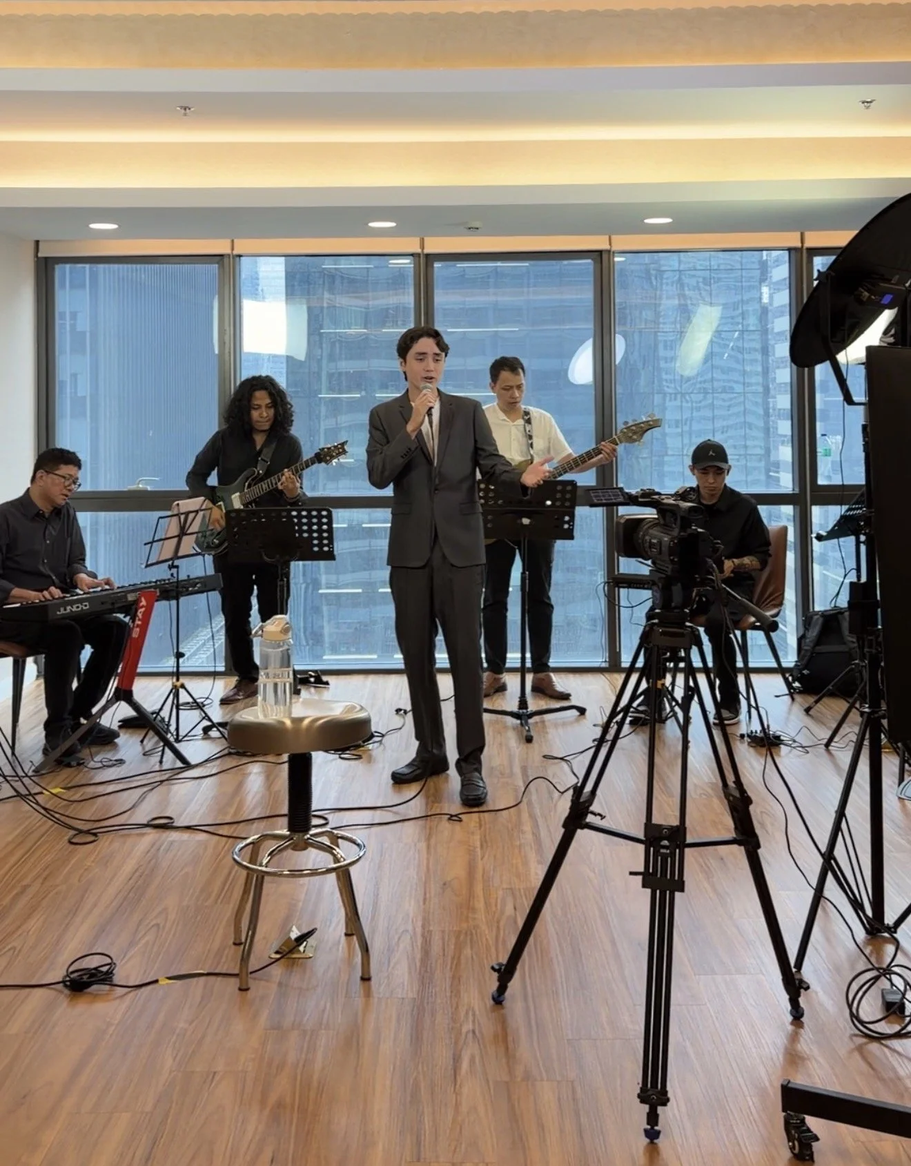 Indoor band performance with five musicians, one singing and four playing instruments, recorded in a room with large windows and cityscape background.