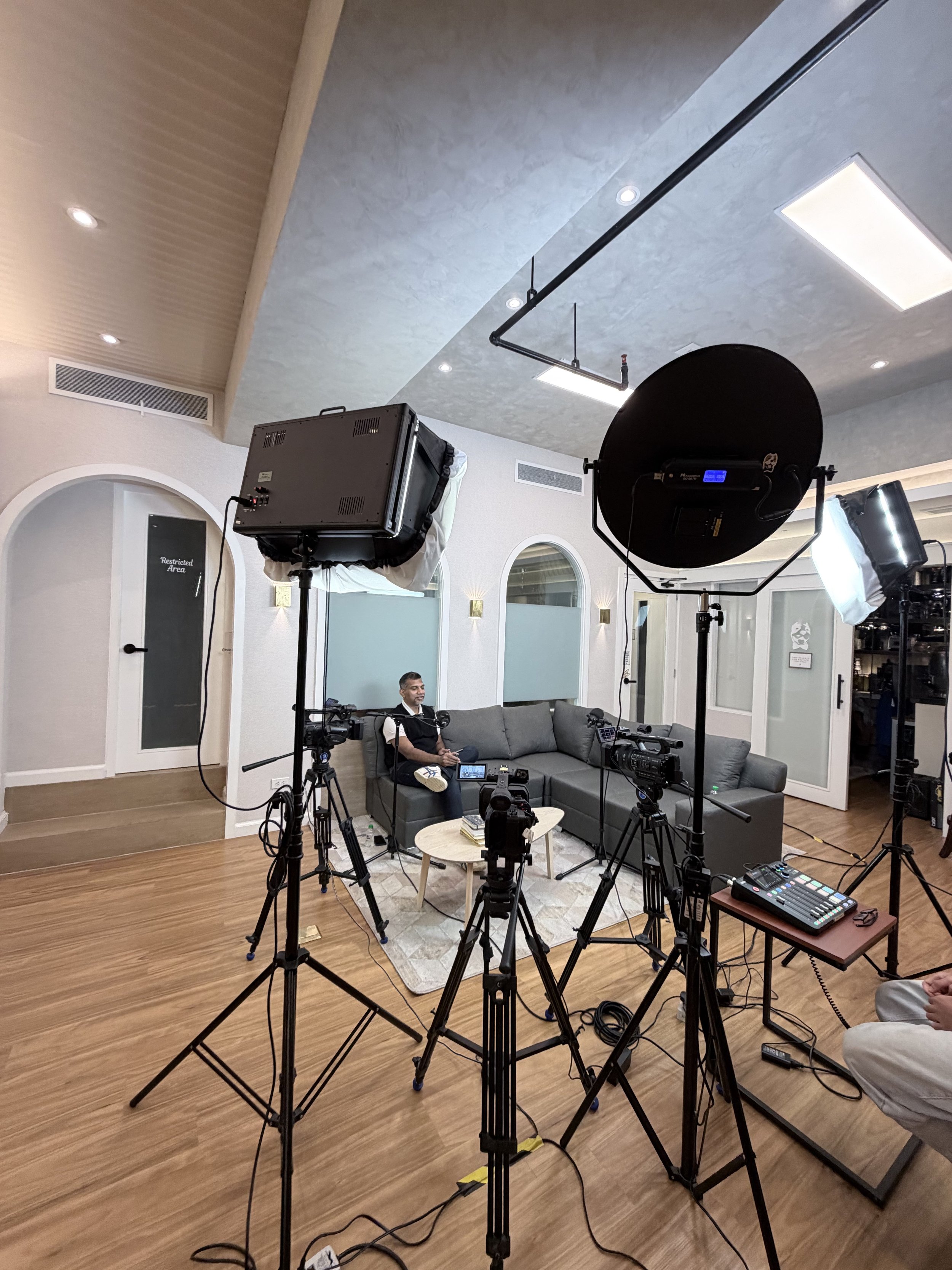A behind-the-scenes view of a recording or live streaming setup in a room. Multiple cameras, lights, and equipment are positioned around a sofa where a man is sitting, seemingly preparing for a video or interview.
