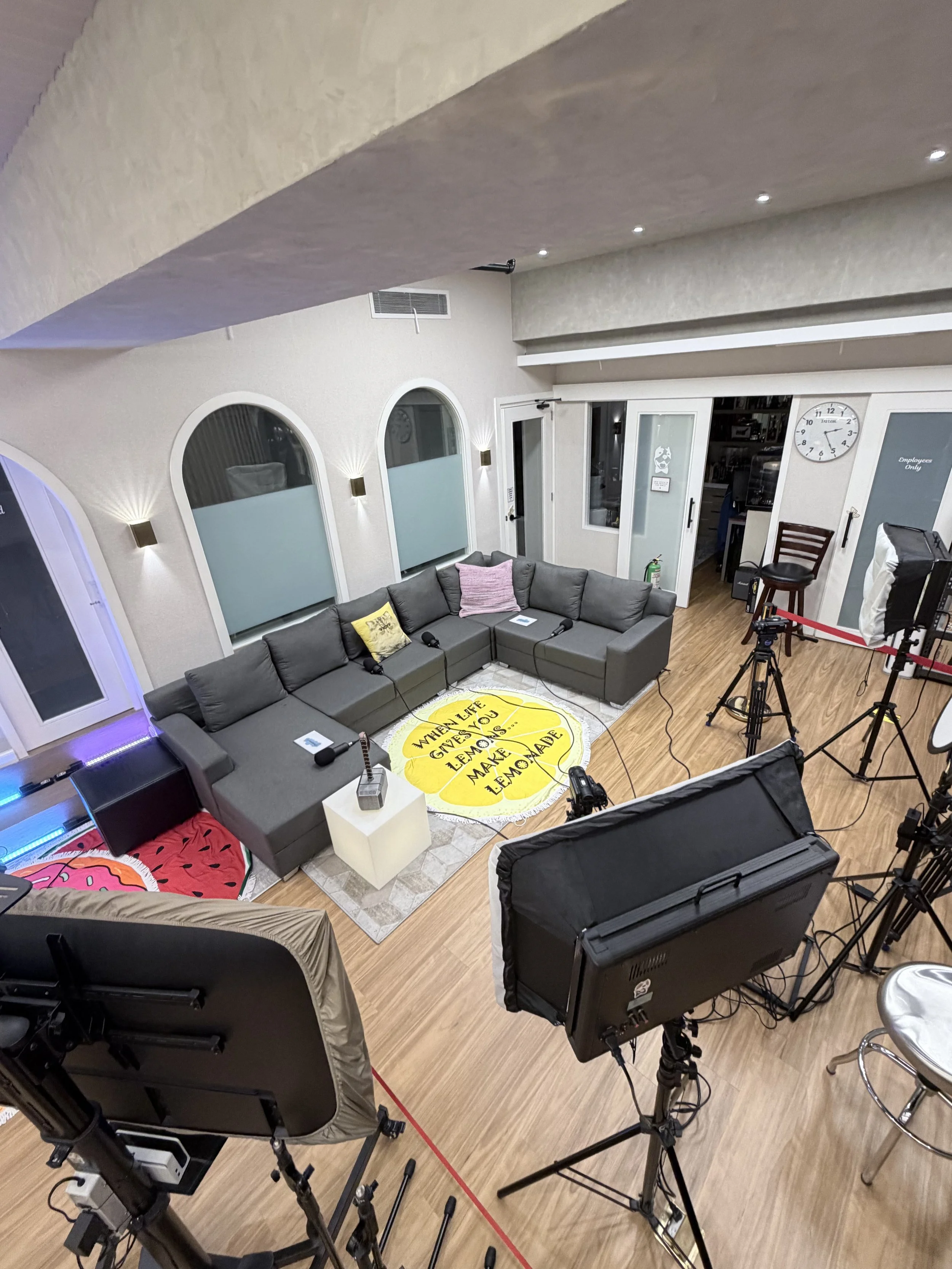 A recording studio or production space with a large gray sectional sofa, a white side table, and various professional photography or filming equipment including lights, tripods, cameras, and a monitor. The room has arched windows, wall-mounted lights