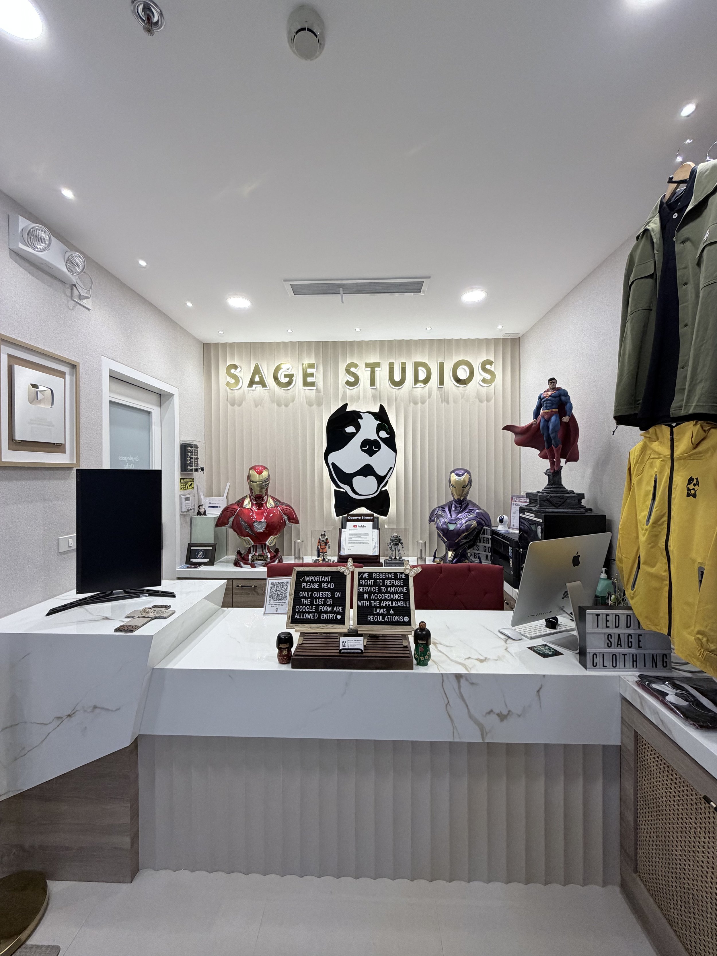 Interior of an office or reception area with a white marble counter, superhero and character statues, framed pictures on the wall, and a sign that reads 'SAGE STUDIOS' on the back wall. There are clothing items hanging on the right side.