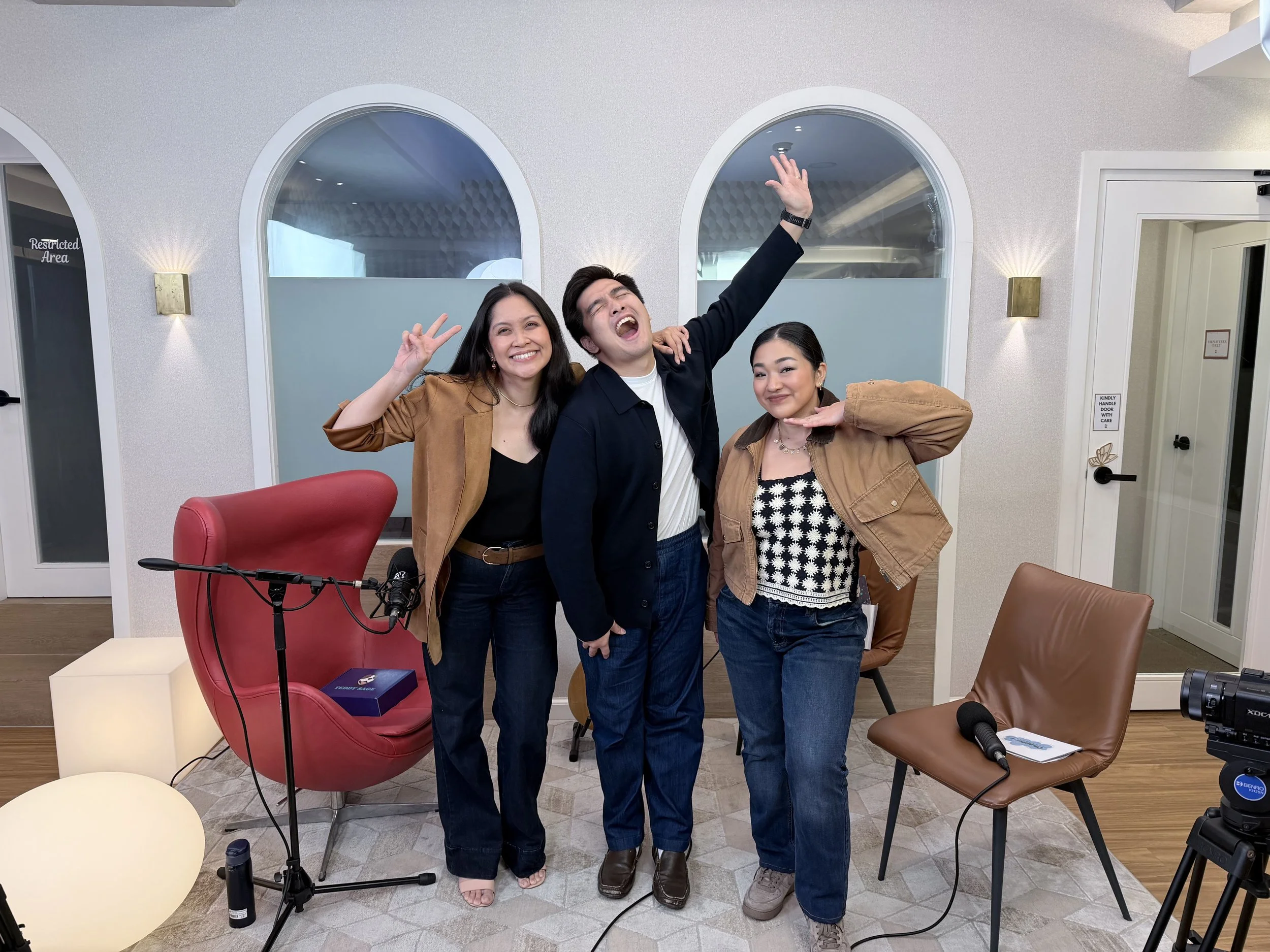 Three people standing behind a table in a room with arched windows, microphones, and chairs. The group is smiling and makes peace signs and a playful pose.
