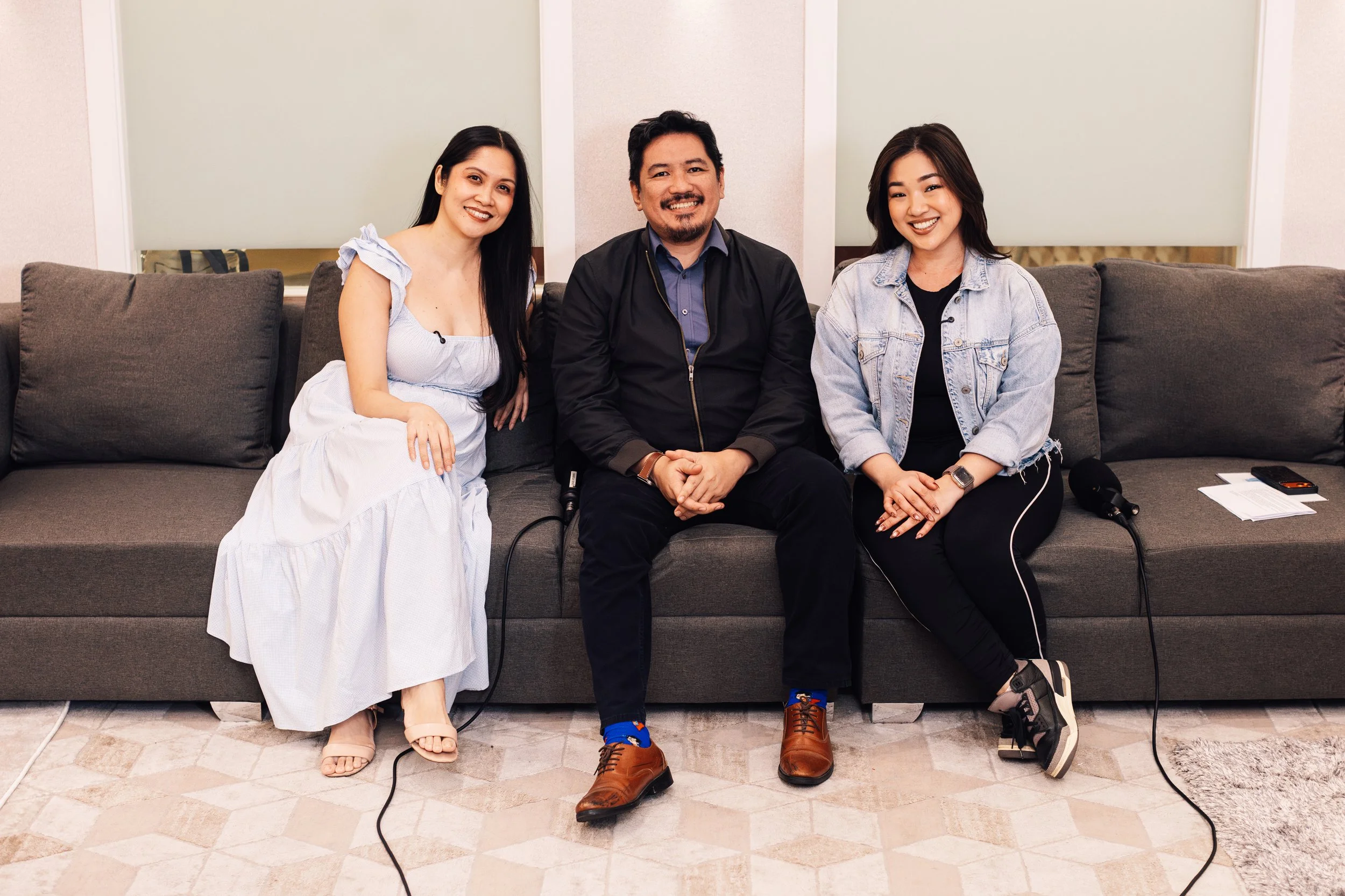 Three people sitting on a gray couch in a room with beige walls. A woman in a white dress is on the left, a man in a black jacket and blue shirt in the center, and a woman in a denim jacket with a black shirt on the right. There is a microphone on th