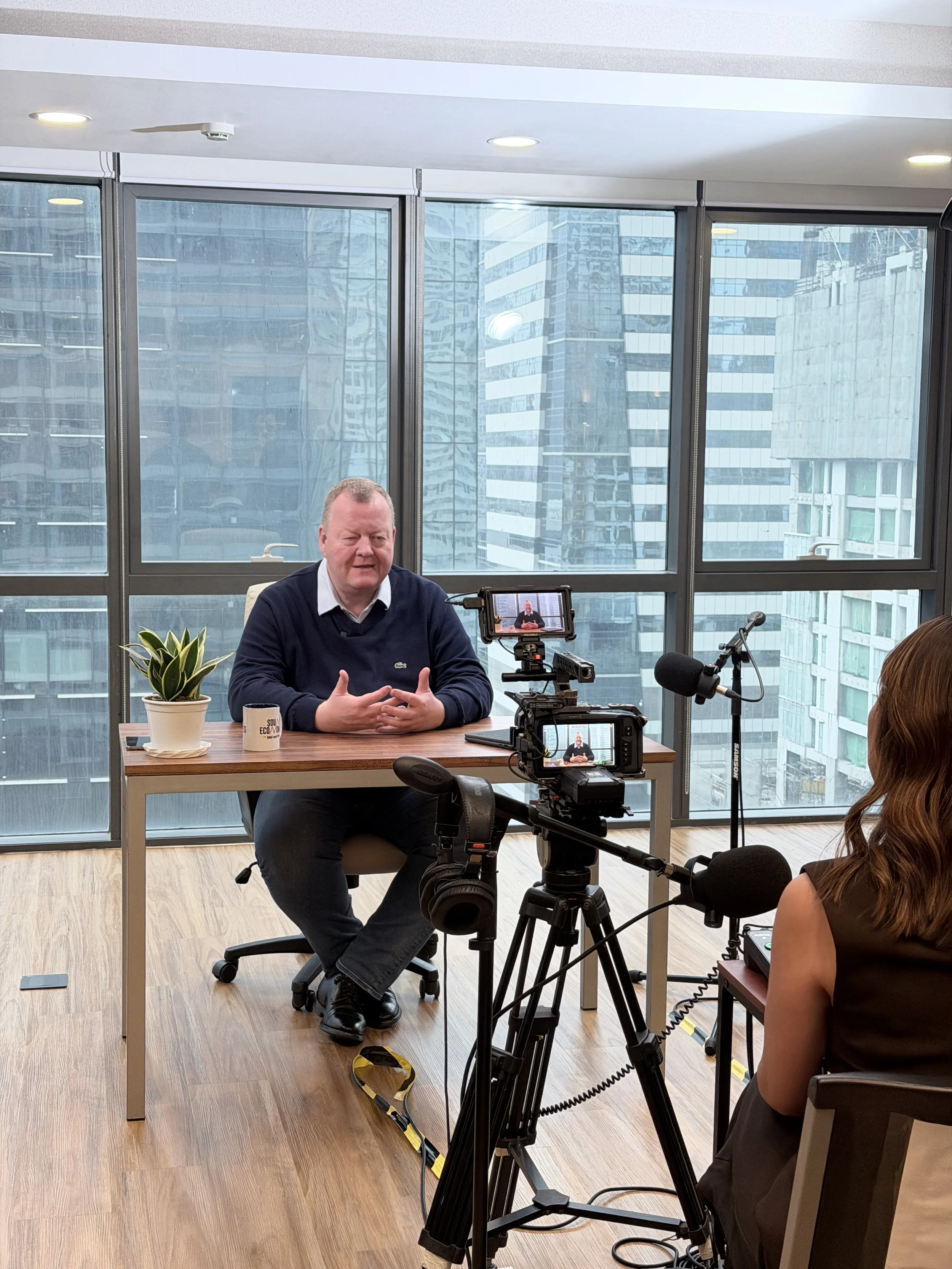 A man sitting at a desk being filmed or recorded by a woman with cameras and microphones in an office setting, with large windows showing tall city buildings outside.