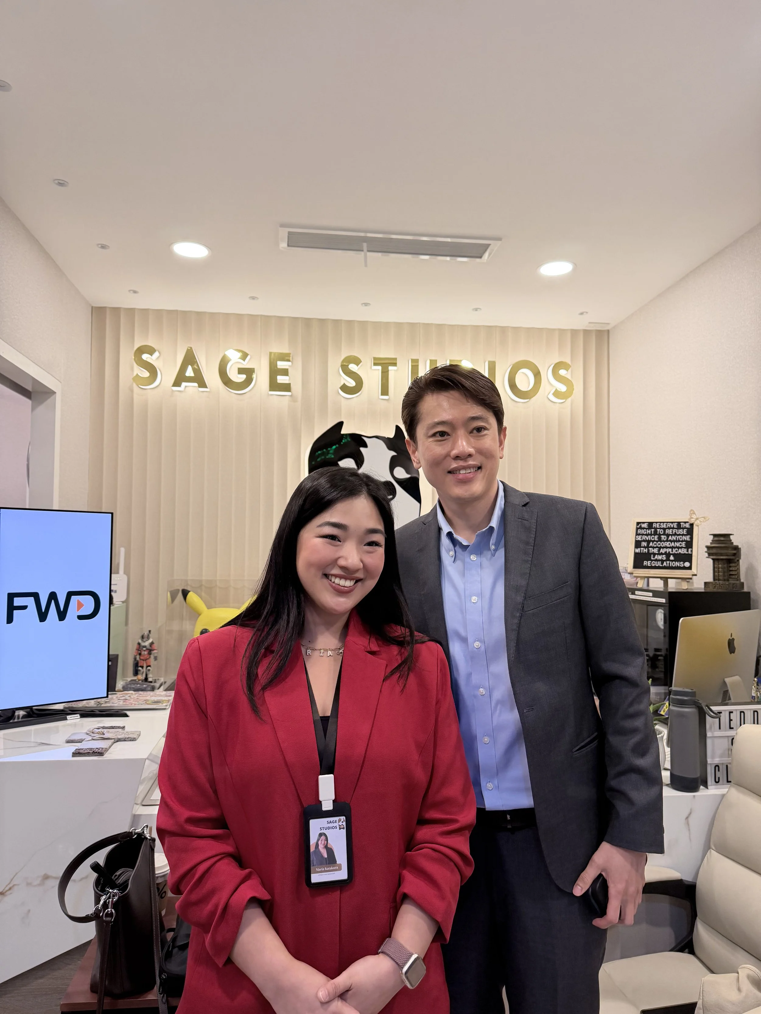 Two people standing in front of a beige wall with the words "SAGE STUDIOS" illuminated on it. The woman on the left is wearing a red blazer and has a badge around her neck. The man on the right is wearing a gray suit and blue shirt.