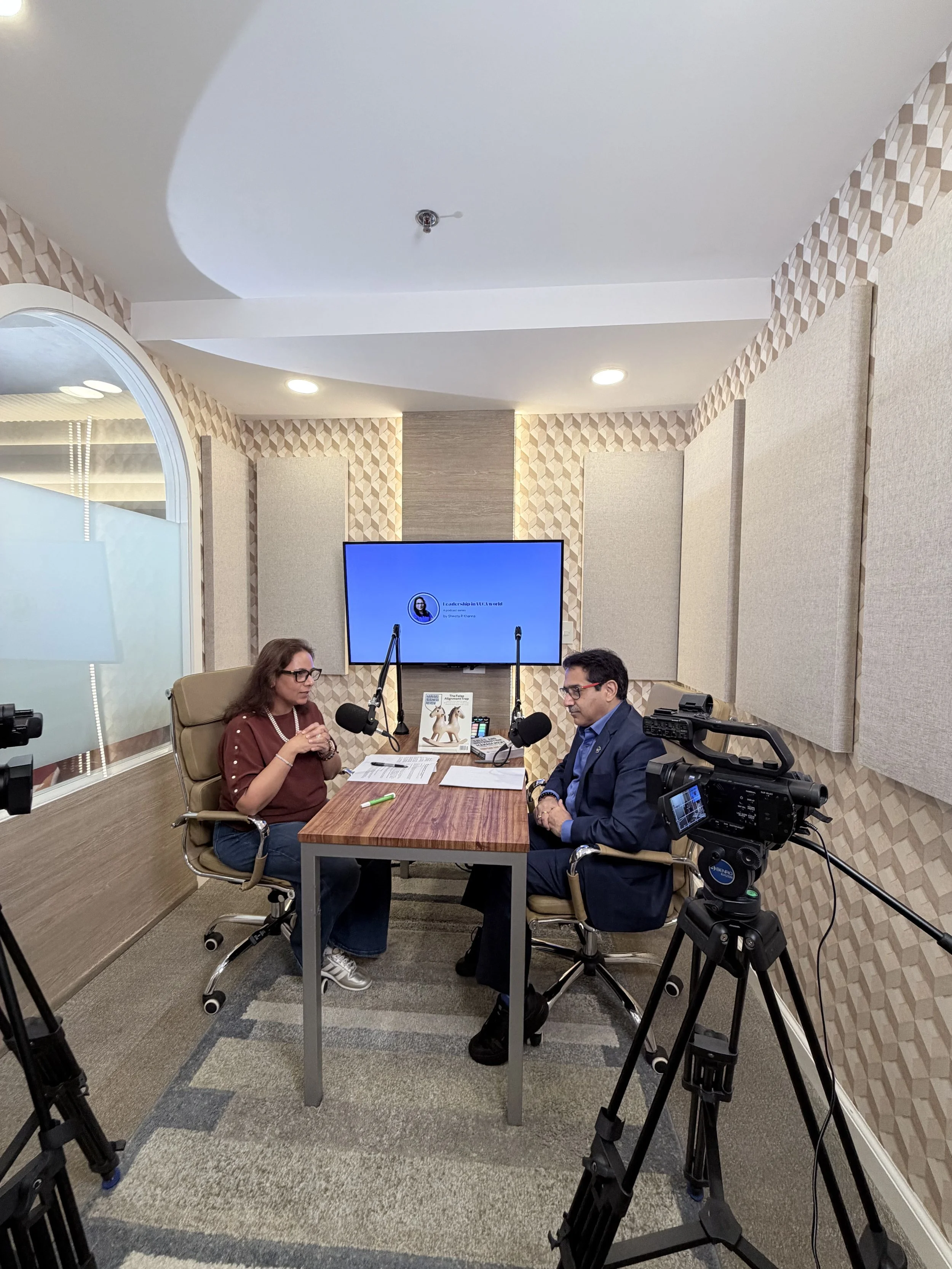 Two people sitting across from each other at a table in a recording studio or podcast room, with cameras and microphones set up for recording.