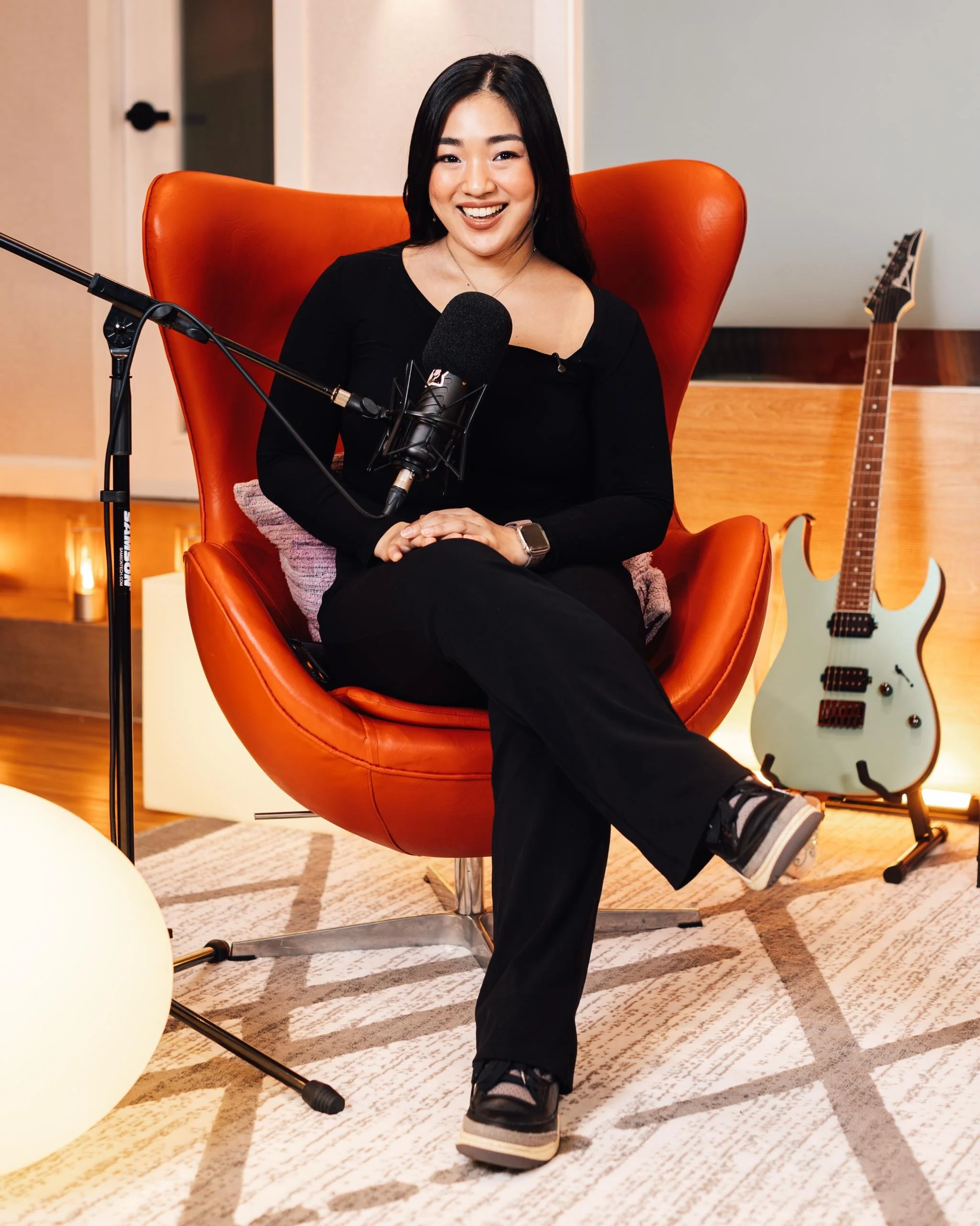 A young woman with dark hair sitting in an orange modern armchair, smiling at the camera in a cozy room with acoustic guitar behind her and a spherical lamp in the foreground.
