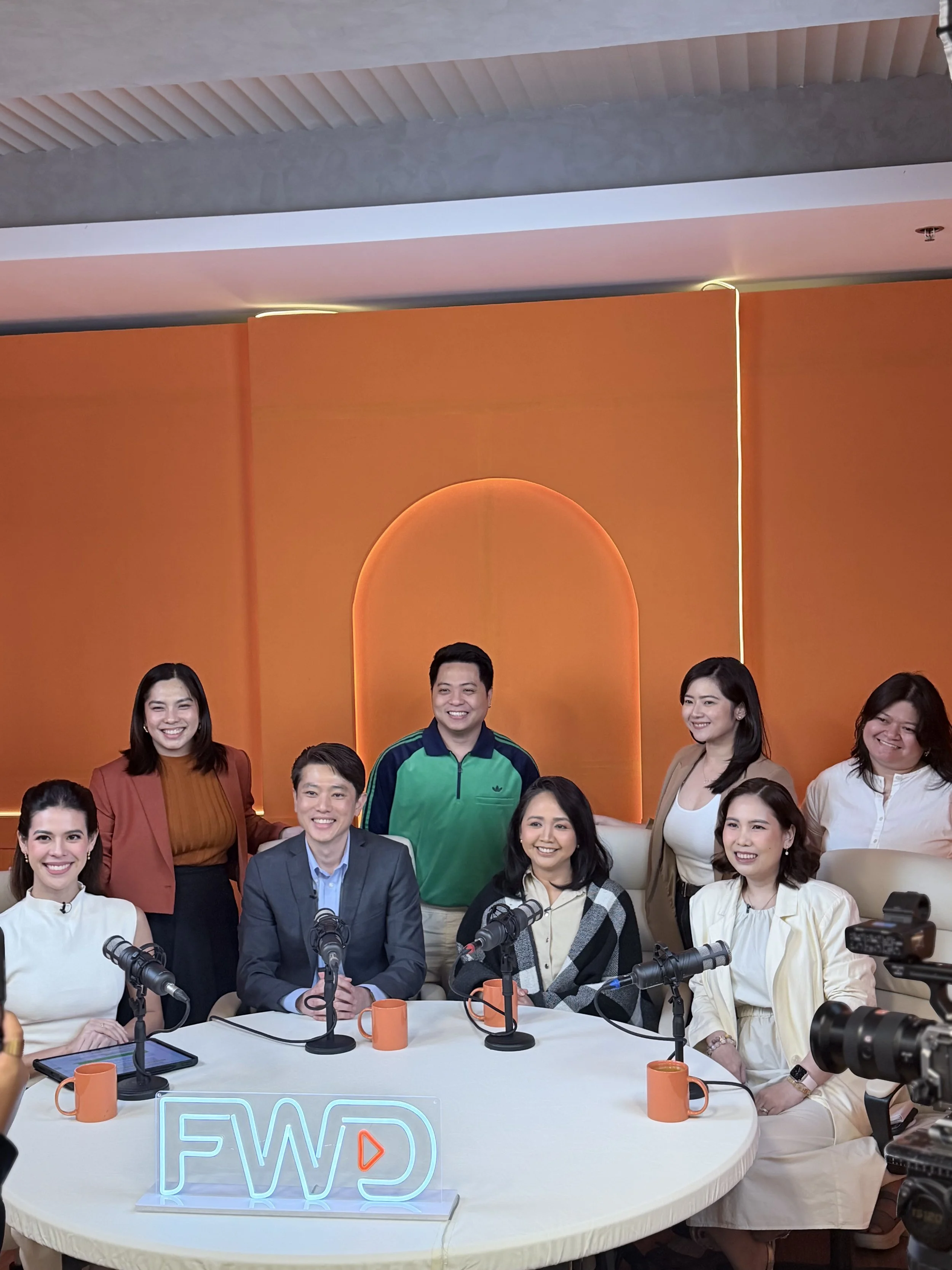 A diverse group of people gathered around a white table with microphones, orange mugs, and electronic devices in a modern, brightly lit studio with an orange background and a neon sign reading 'FWD'.