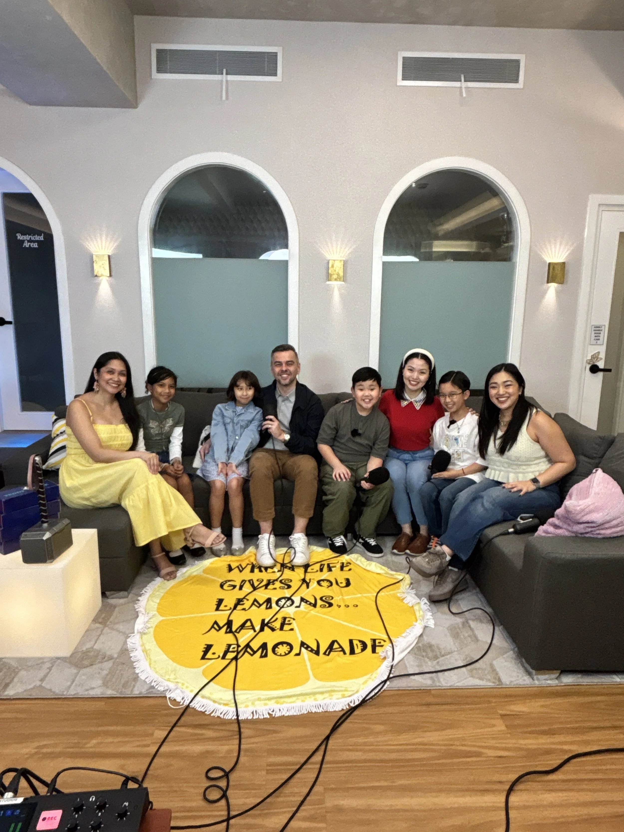 A group of eight people sitting on a sofa, smiling at the camera. They are surrounded by a microphone, audio equipment, and a large lemon-shaped banner on the floor with black text that says, 'Who will give you lemons... make lemonade.' The backgroun