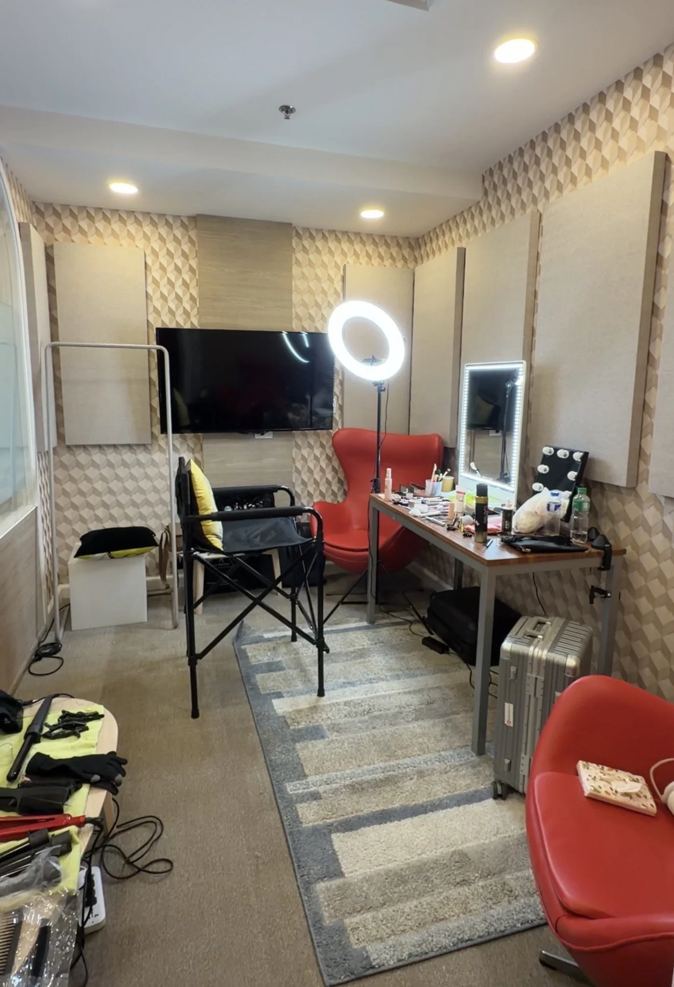 A room with beige patterned wallpaper, a flat-screen TV on the wall, a vanity mirror with lights, a red chair, a table cluttered with makeup and tools, two additional chairs, a metal case, and a small wooden table with tools and supplies.