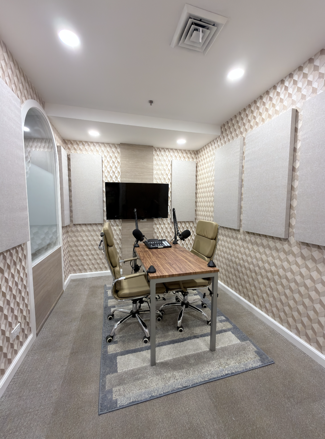 Recording studio with a wooden table, four beige chairs, a large flat-screen TV on a wall with beige acoustic panels, and two microphones.