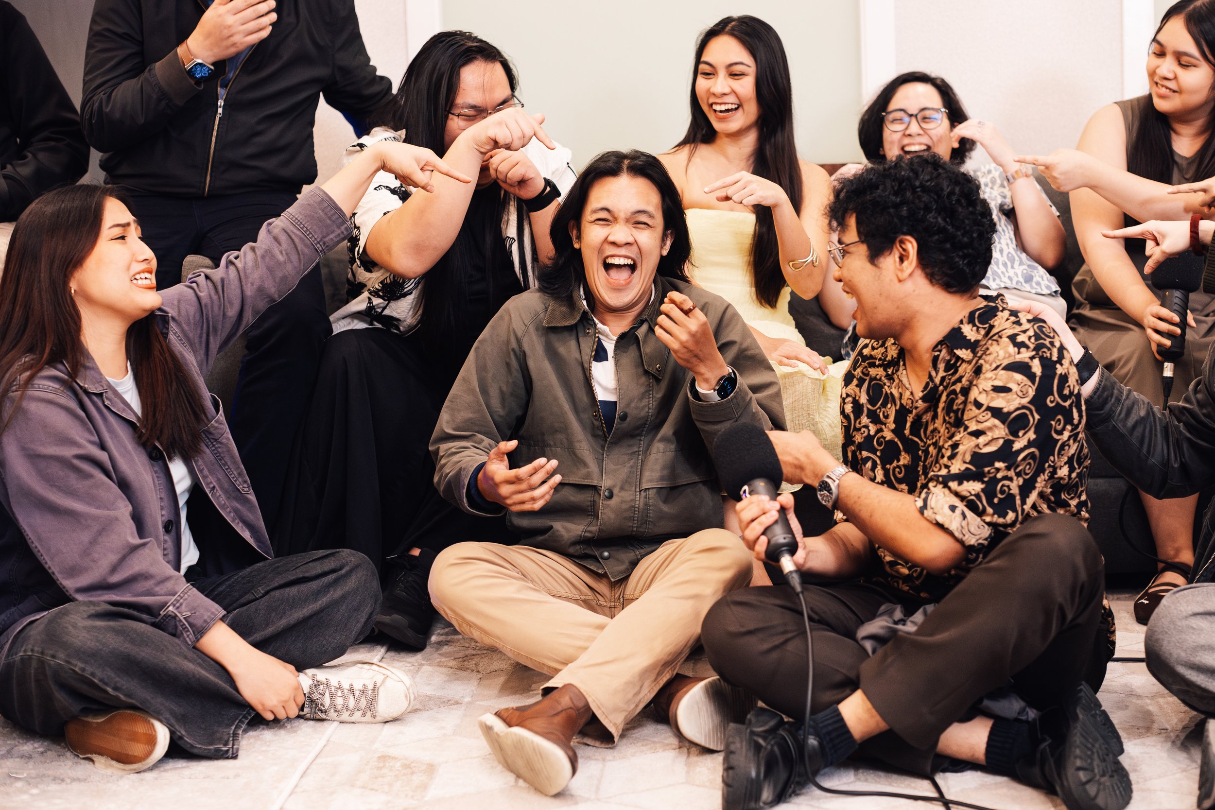 A group of people, mostly women, sitting on the floor in a circle, laughing and smiling. Some are pointing at each other, and one person is holding a microphone, indicating a lively and joyful gathering.