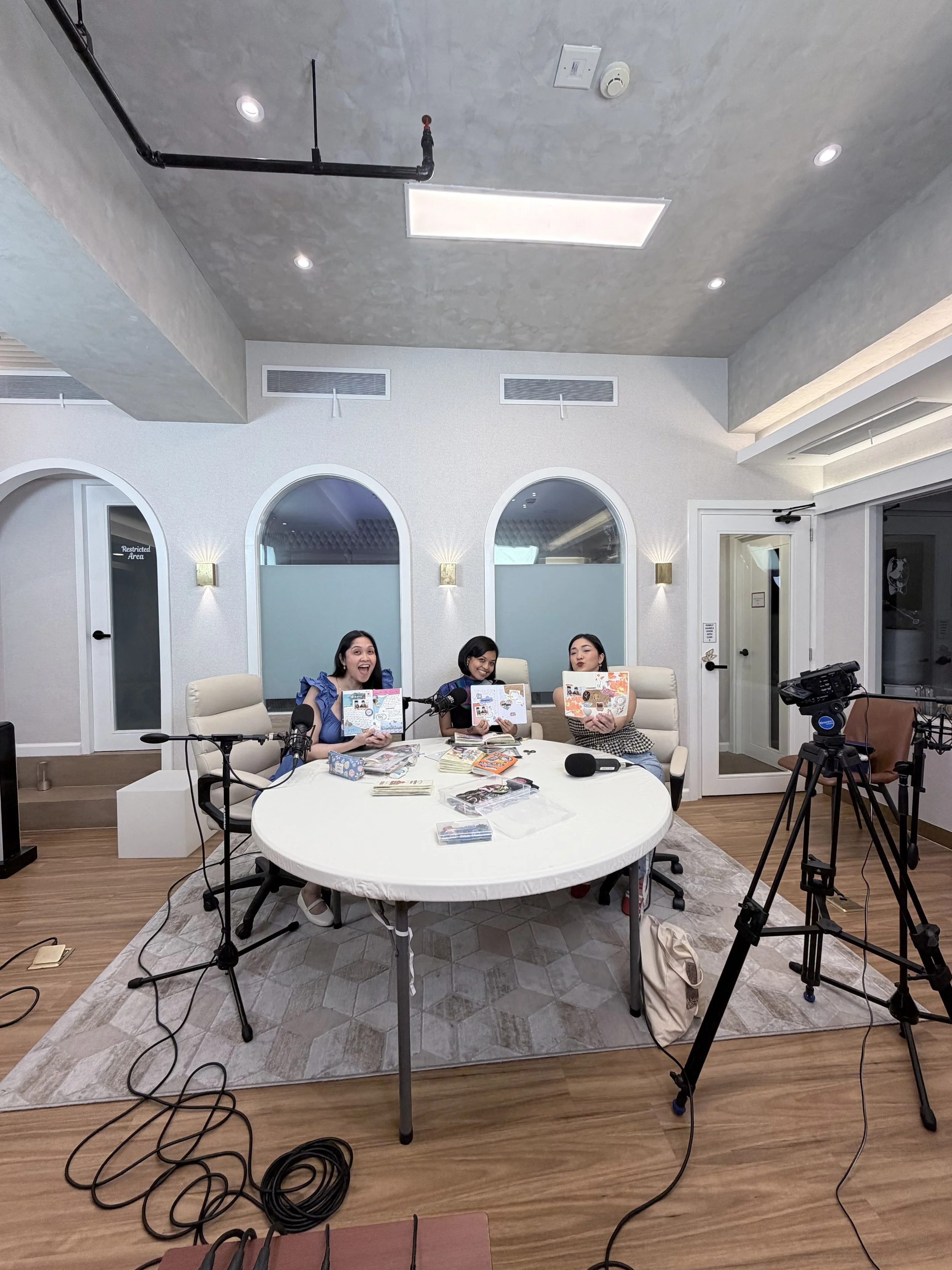 Three women sitting at a table during a recording or podcast session. They are holding magazines or books and are surrounded by microphones, camera, and other recording equipment in a well-lit room with white walls and arched windows.