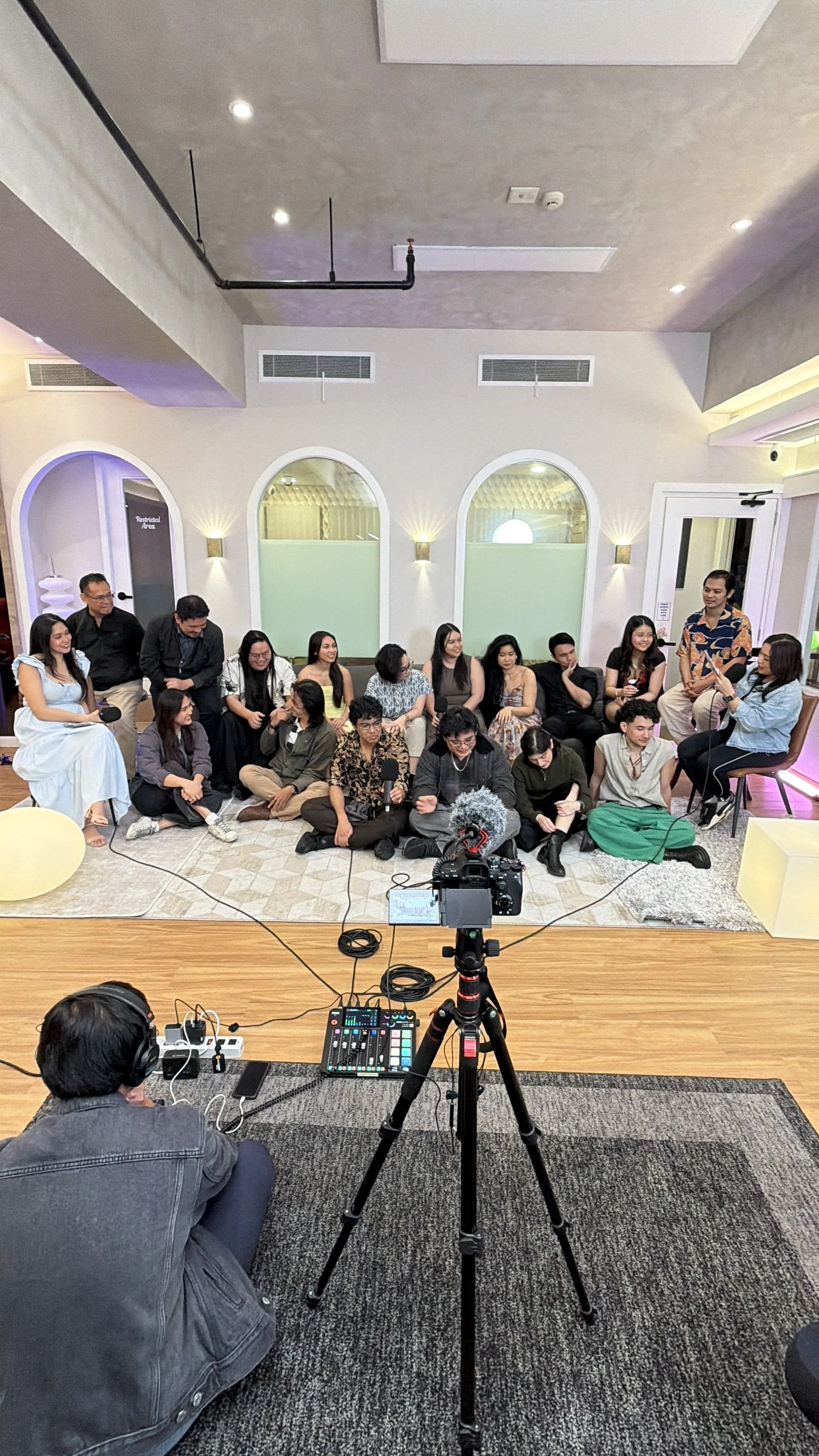A group of people sitting and standing in a room, participating in an event or interview, with camera and audio equipment in the foreground.