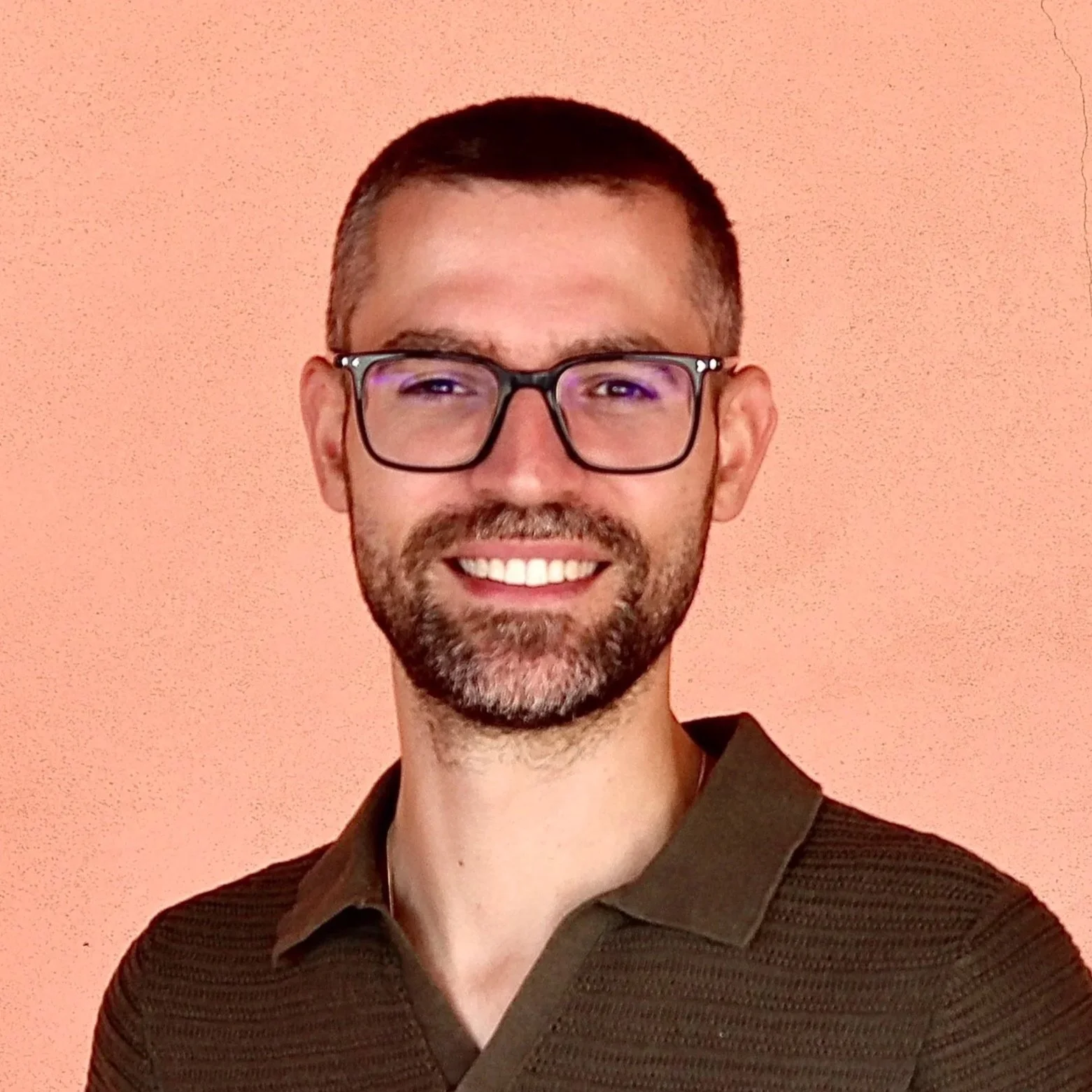Homem com óculos, barba e sorriso, usando camisa polo em fundo rosa pálido.
