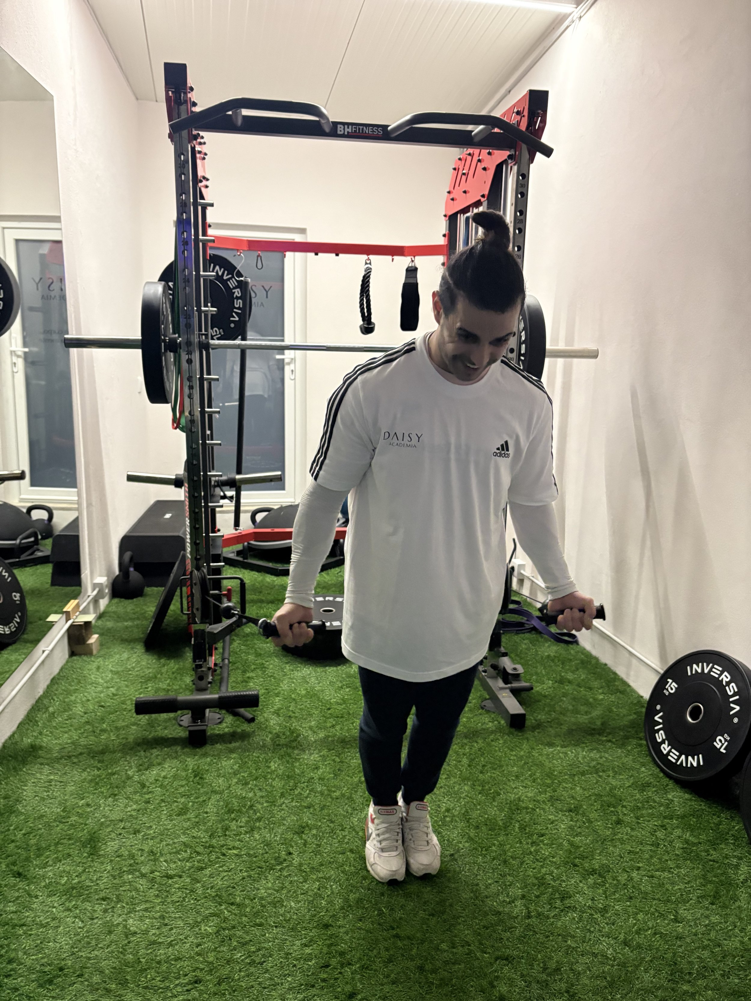Homem realizando levantamento de peso com halteres em uma academia, com equipamento de musculação ao fundo e piso de grama artificial.