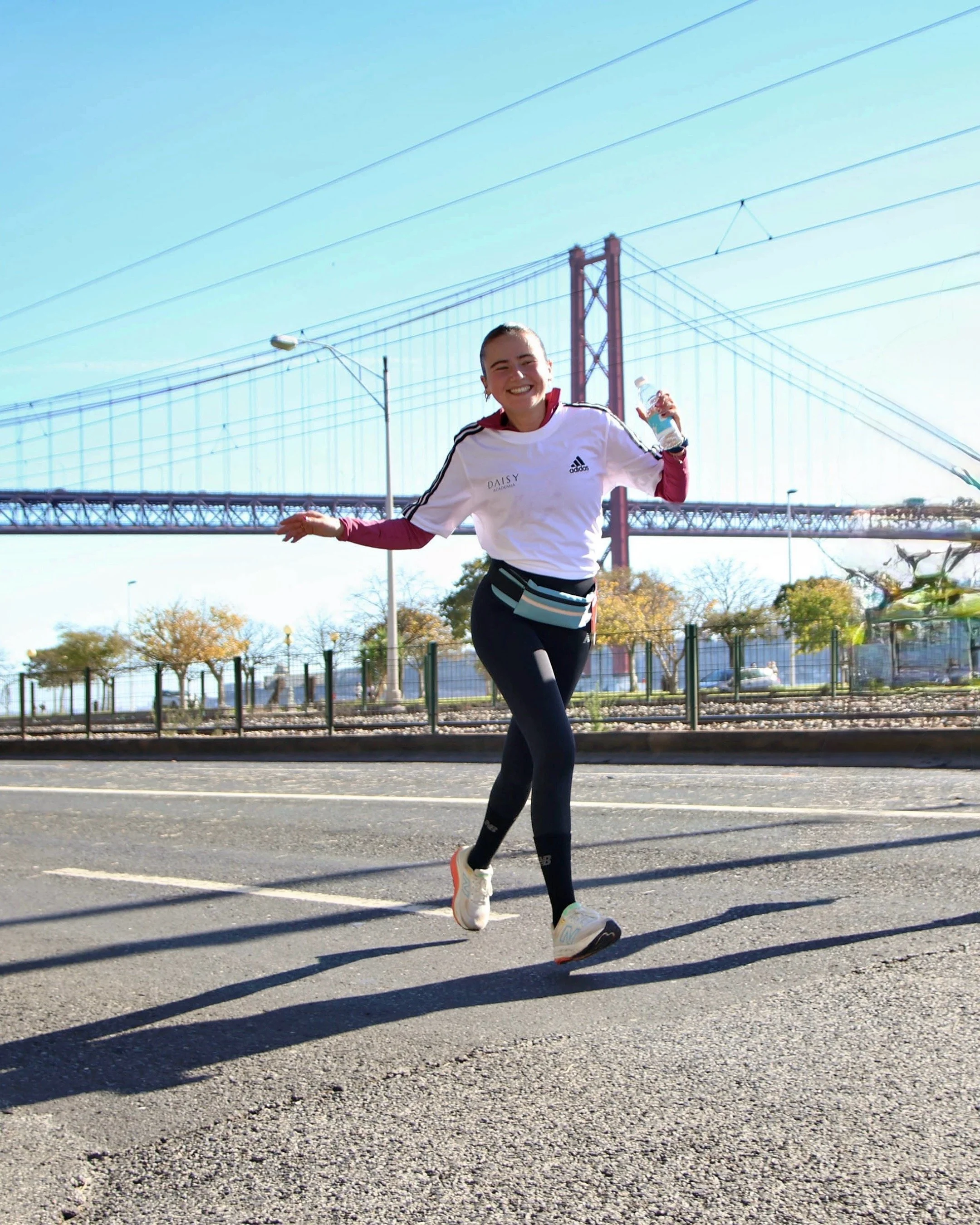 Mulher sorridente correndo na rua com uma ponte ao fundo, vestindo roupas esportivas