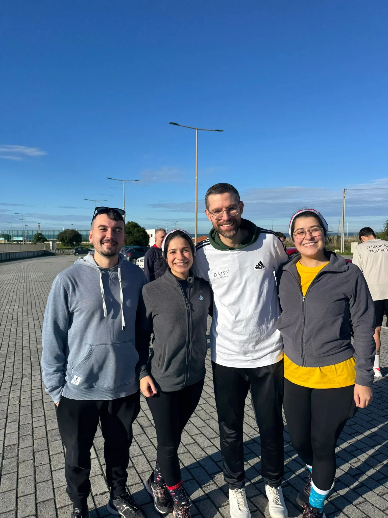 Grupo de quatro pessoas sorrindo ao ar livre em um dia ensolarado, vestindo roupas esportivas, com céu claro e postes de luz ao fundo.