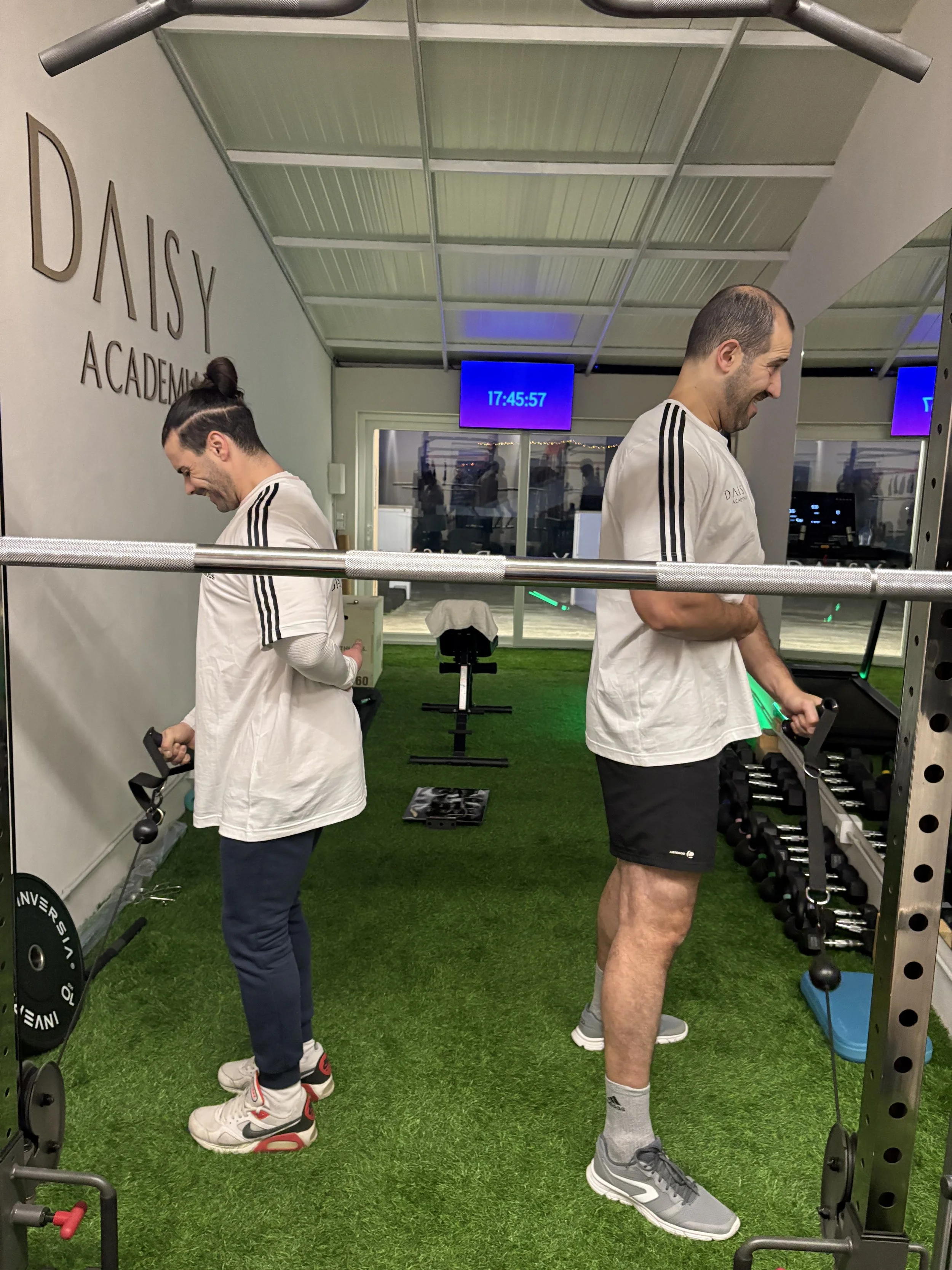 Dois homens treinando com equipamentos de musculação na academia Daisy, com piso de grama artificial, um deles ao lado de um rack de pesos e o outro perto de uma esteira, ambos usando camisetas brancas e expressando diversão.