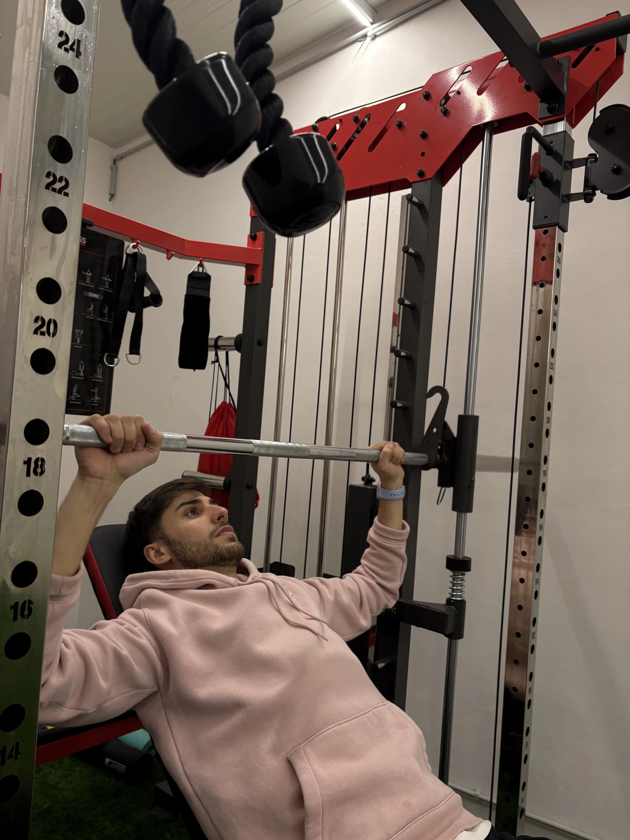 Homem deitado em banco de supino em academia, segurando uma barra com as duas mãos enquanto realiza exercício de supino.