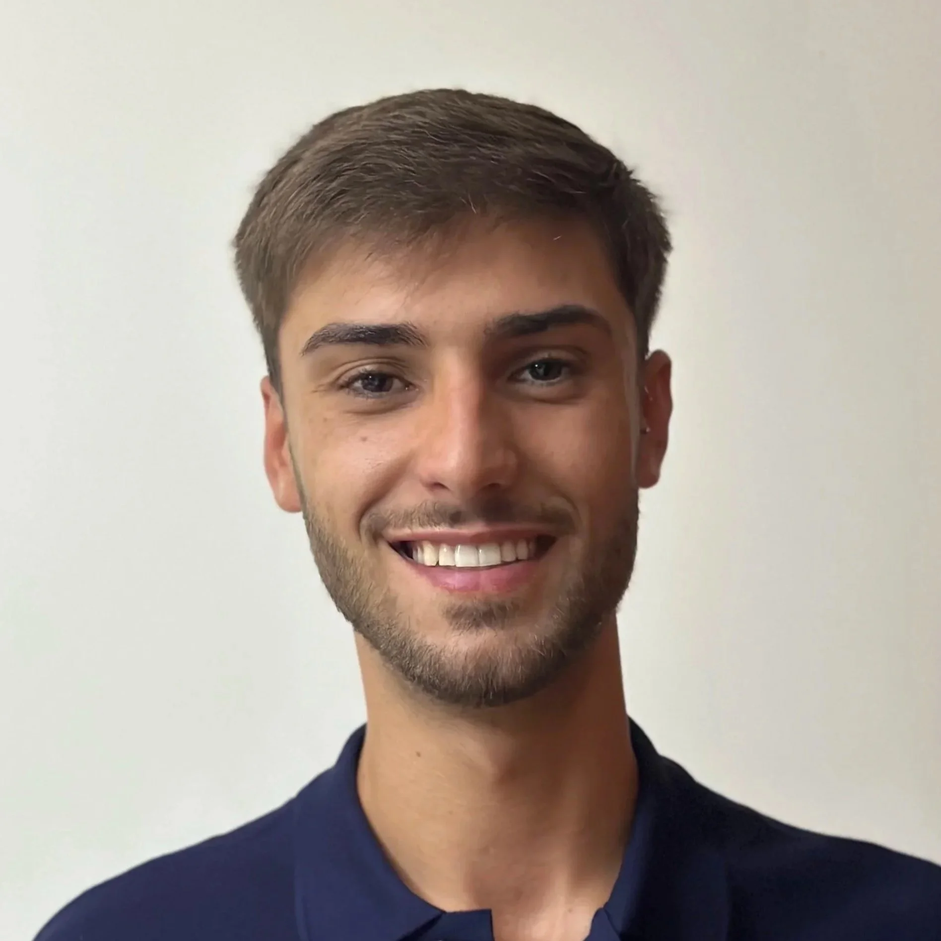 Retrato de um jovem sorridente com pele clara, cabelo castanho, barba por fazer, usando uma blusa escura e fundo branco.