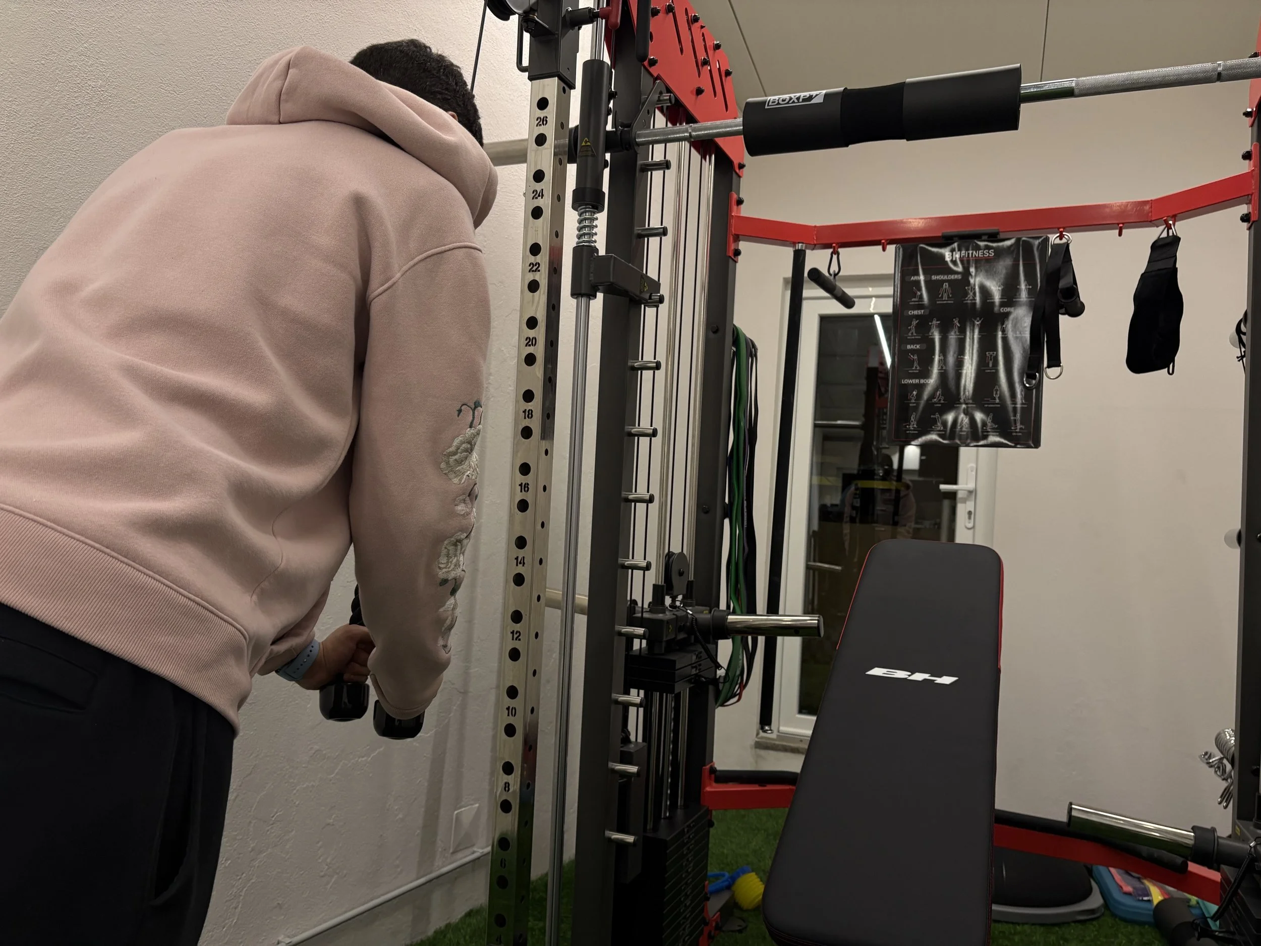 Pessoa de camiseta rosa treinando com halteres em uma academia, perto de uma estrutura de barras de exercício com acessórios negros e uma bicicleta de sombra no fundo.